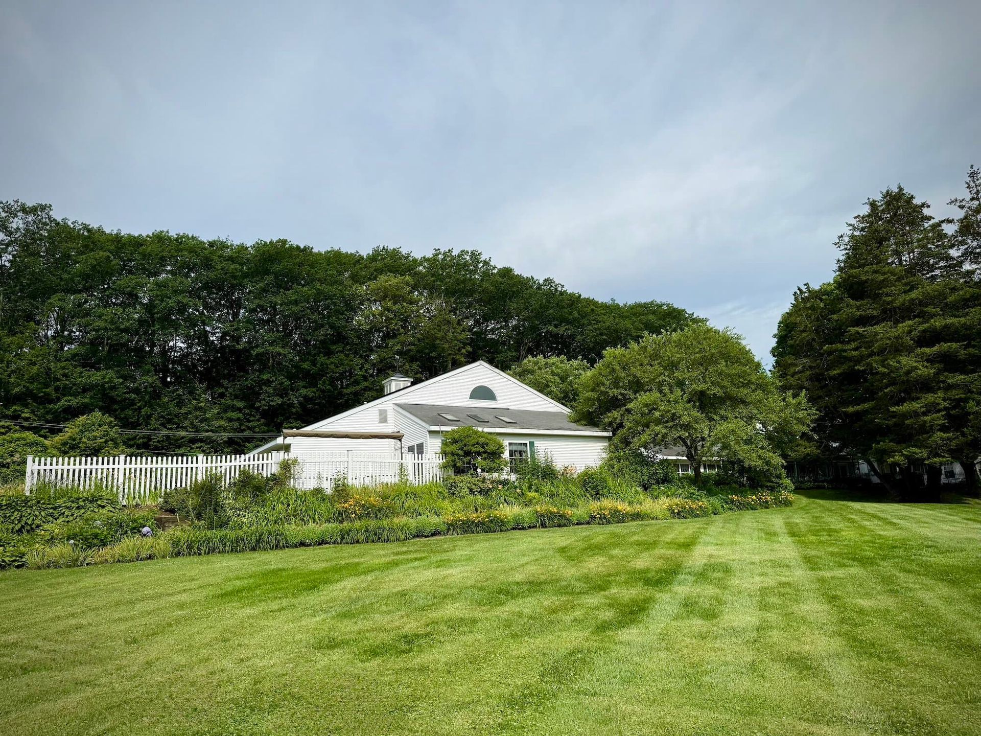 White house with a green lawn, garden, and a treeline under a cloudy sky.