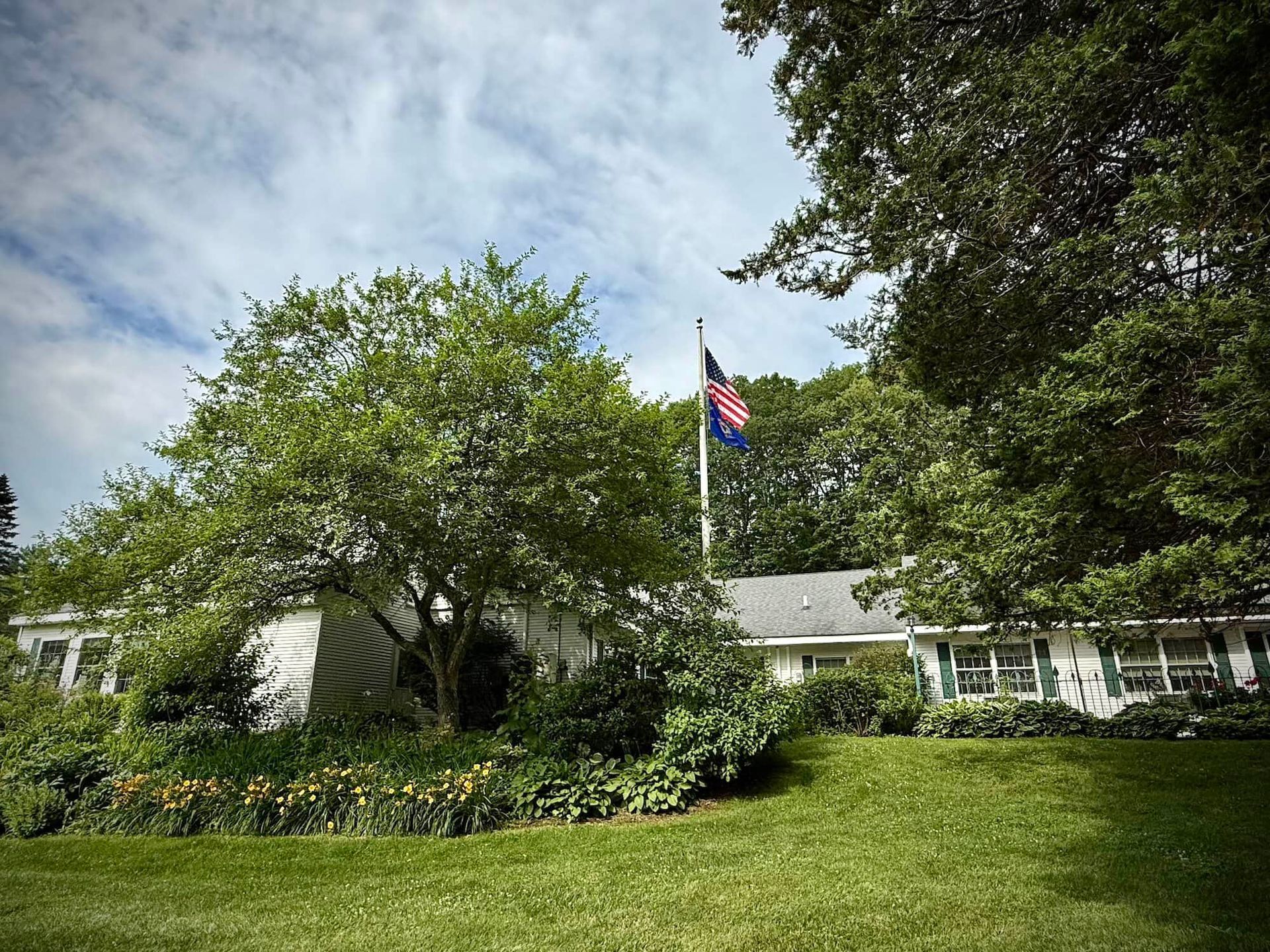White building with American flag flying, trees, and lawn under a cloudy sky.