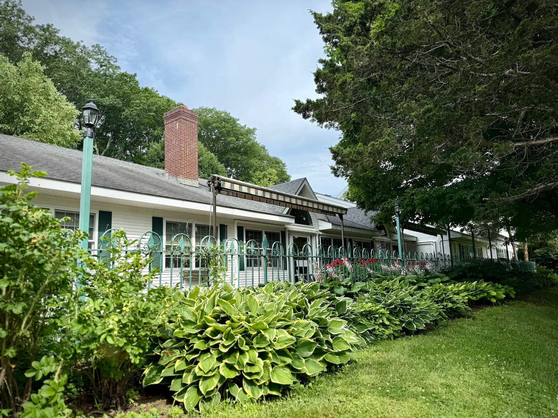 White building with a brick chimney, long row of windows, green trim, and lush greenery.