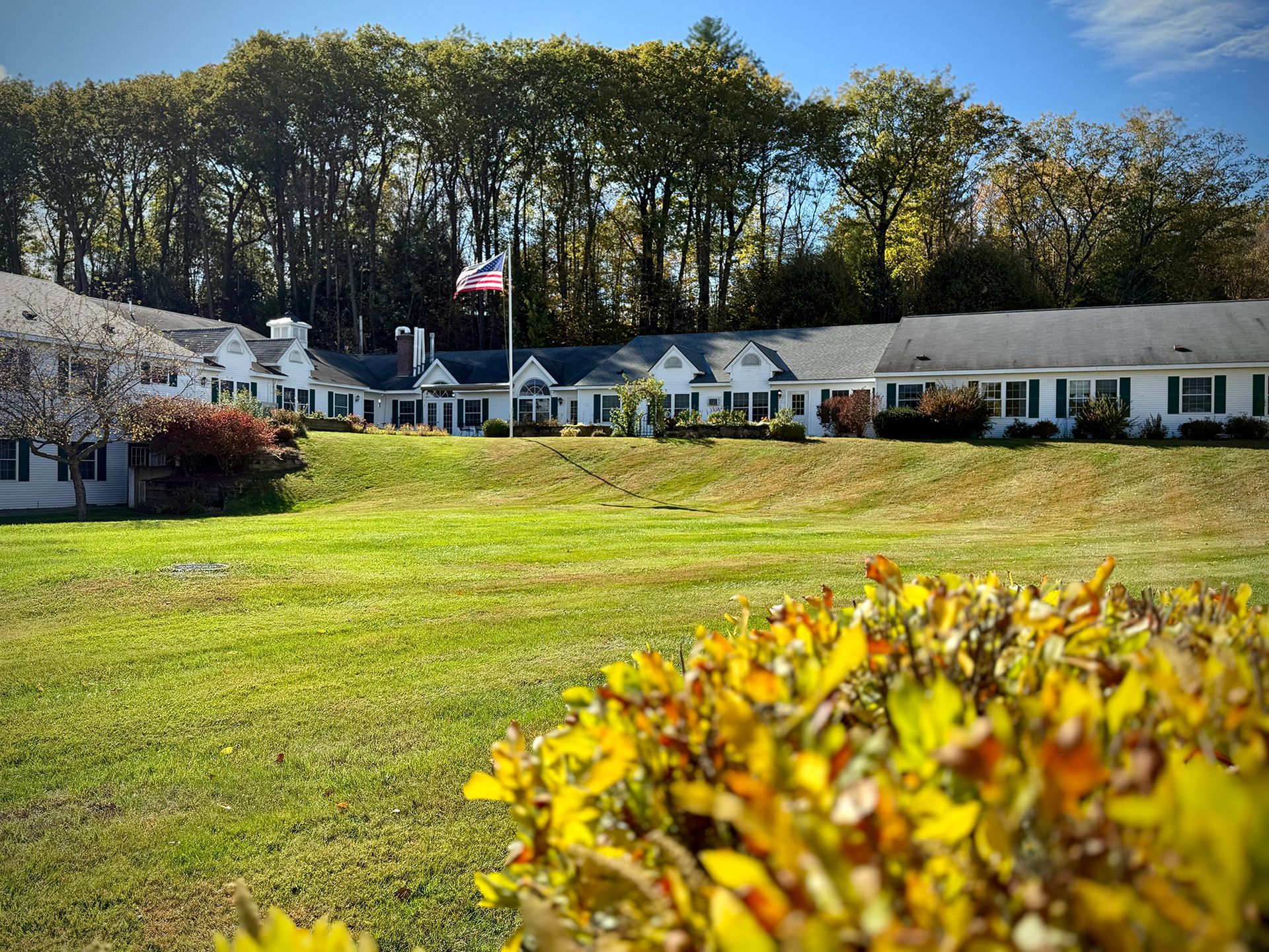 Long, white building with black shutters, American flag, and green lawn, surrounded by trees.