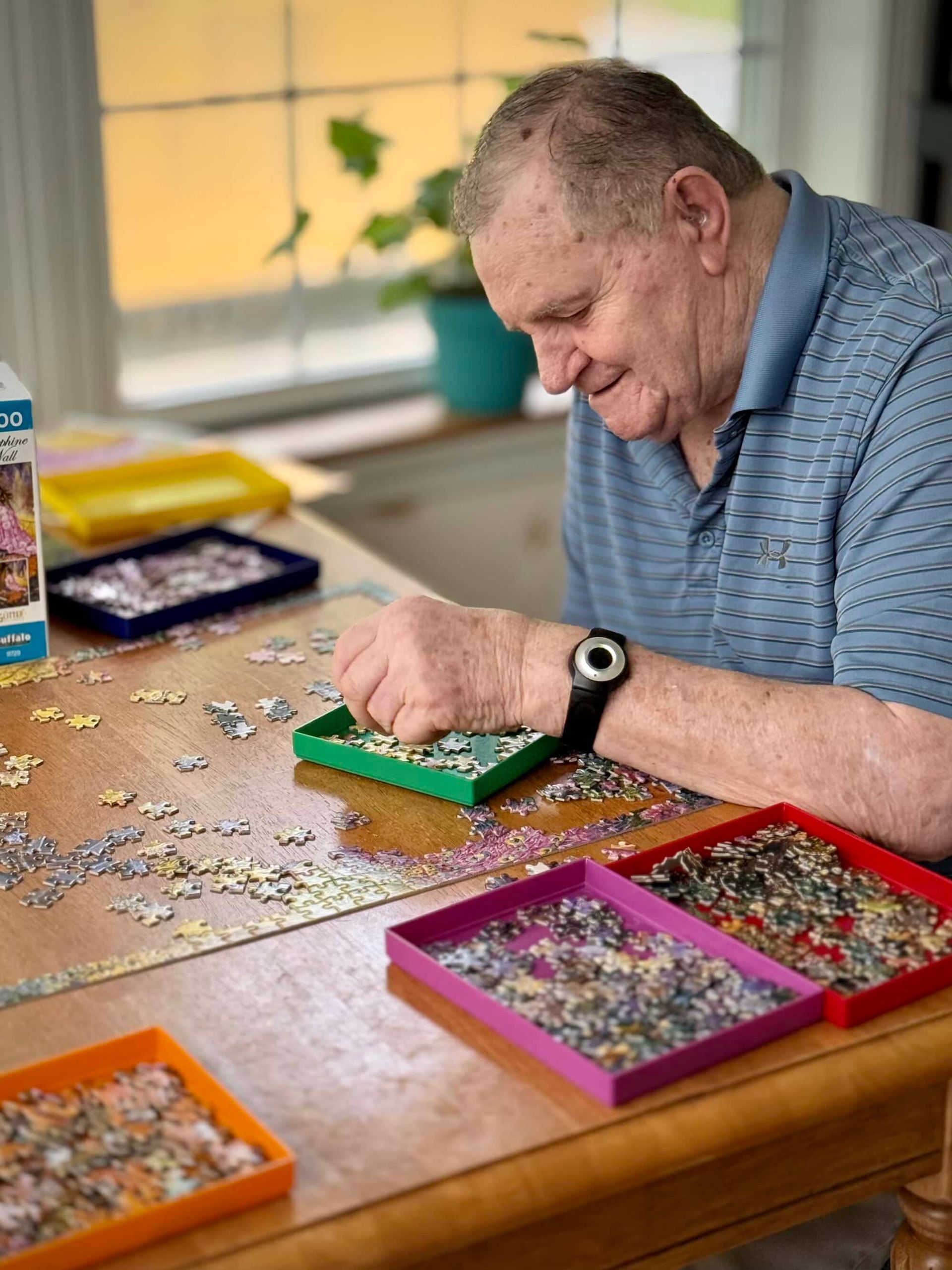 Man seated at a table doing a jigsaw puzzle, using trays for sorting. Smiling, indoors.