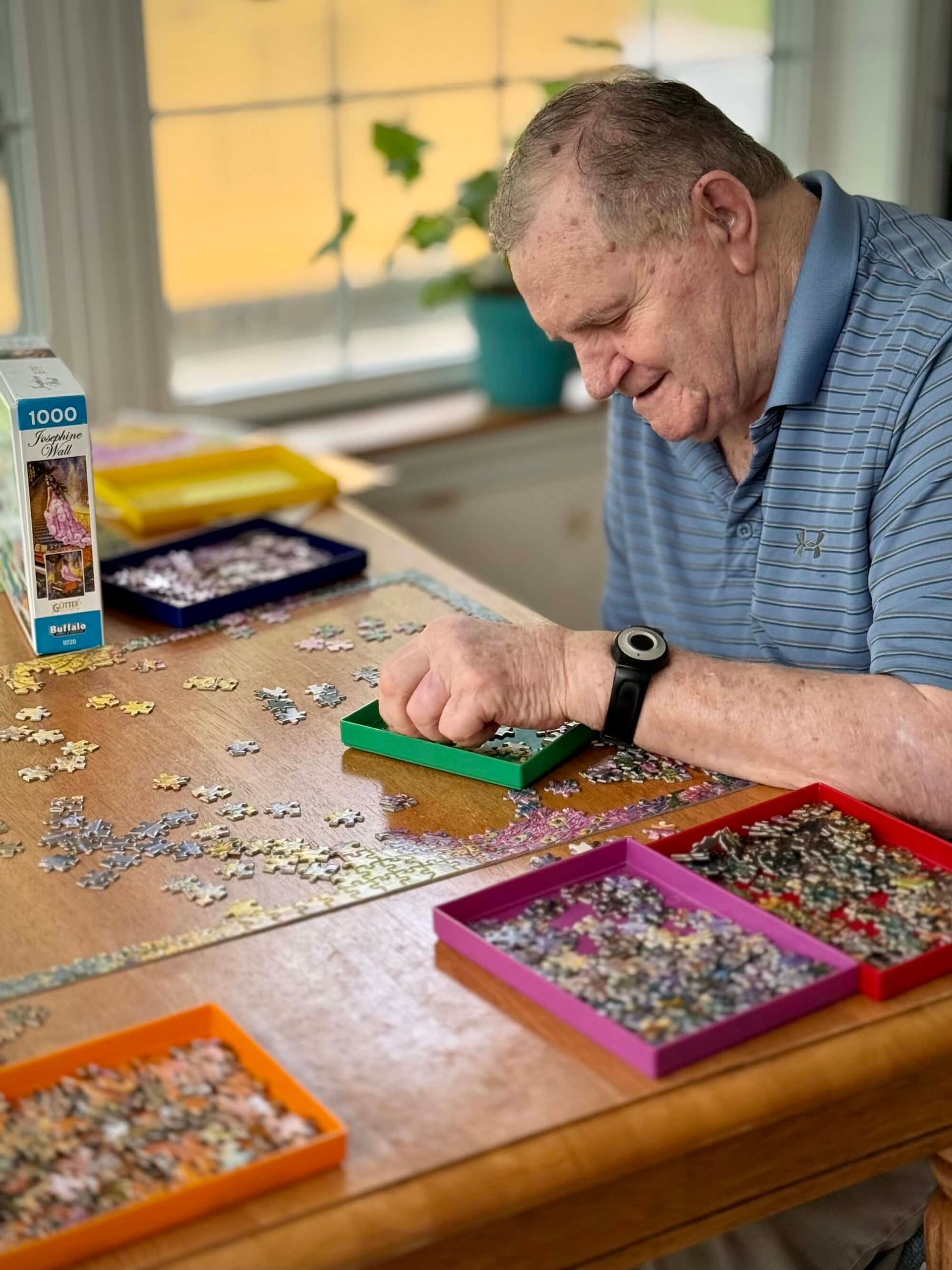 Older adult smiles while working on a jigsaw puzzle at a table, using colored trays to sort pieces.
