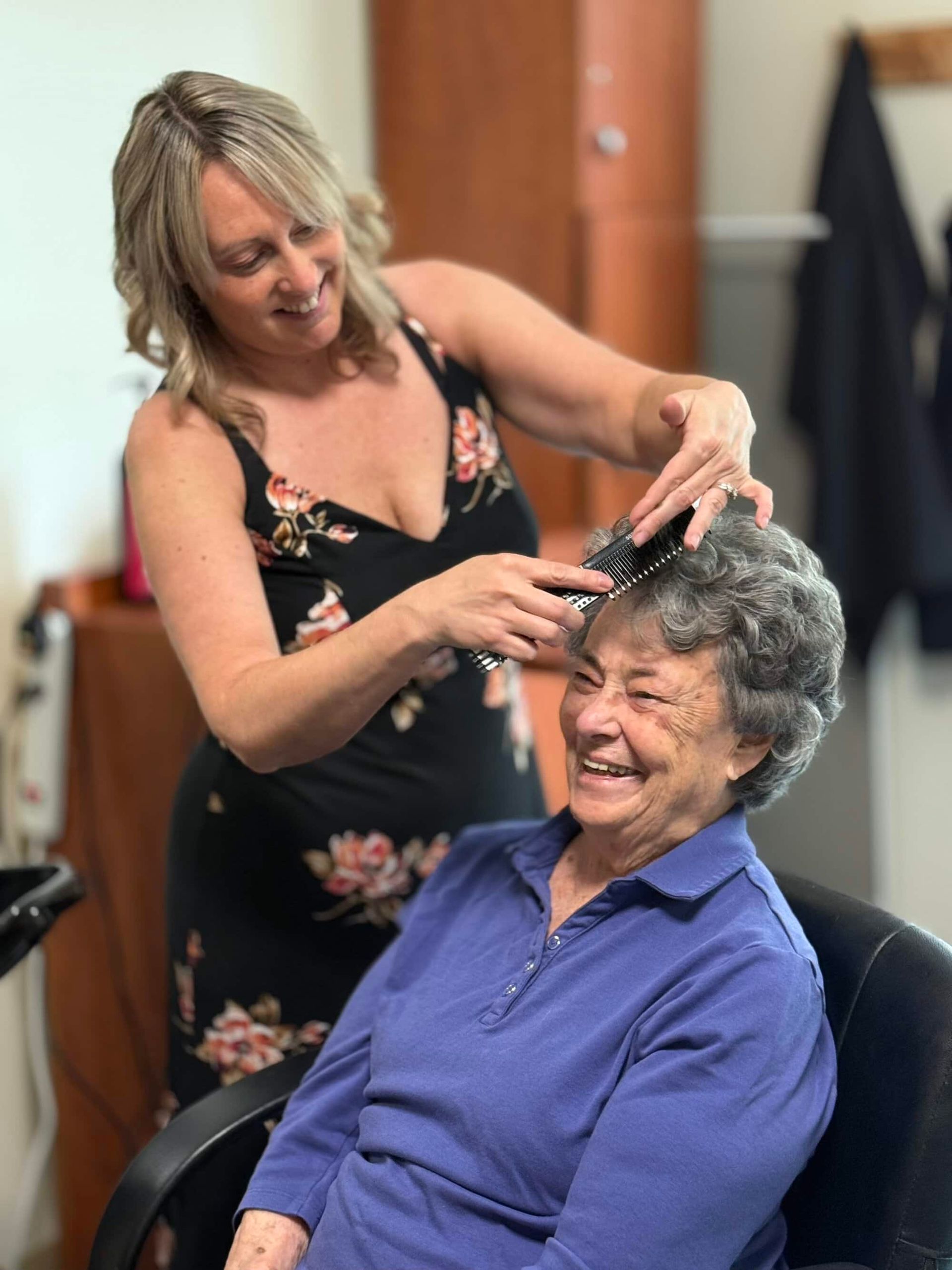 Woman brushing older woman's hair in a salon; both smiling. The younger woman wears a black floral dress.