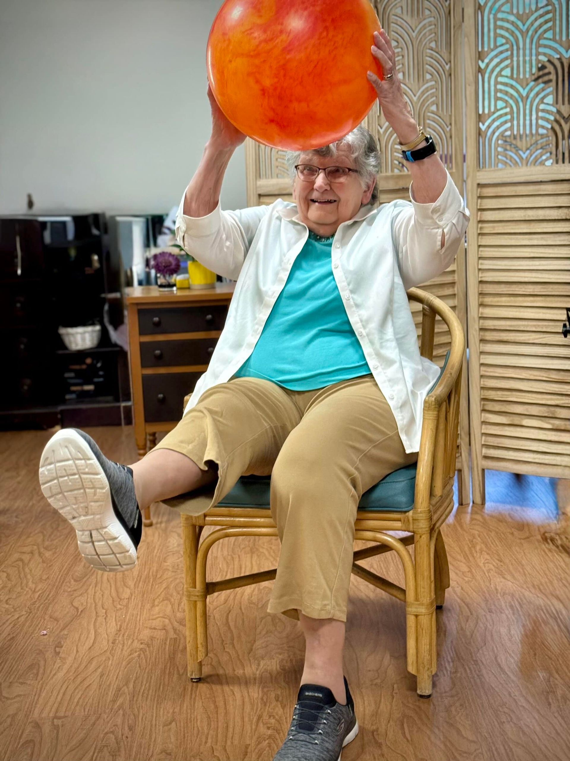Woman seated in a chair, holding a large orange ball overhead, lifting one leg.