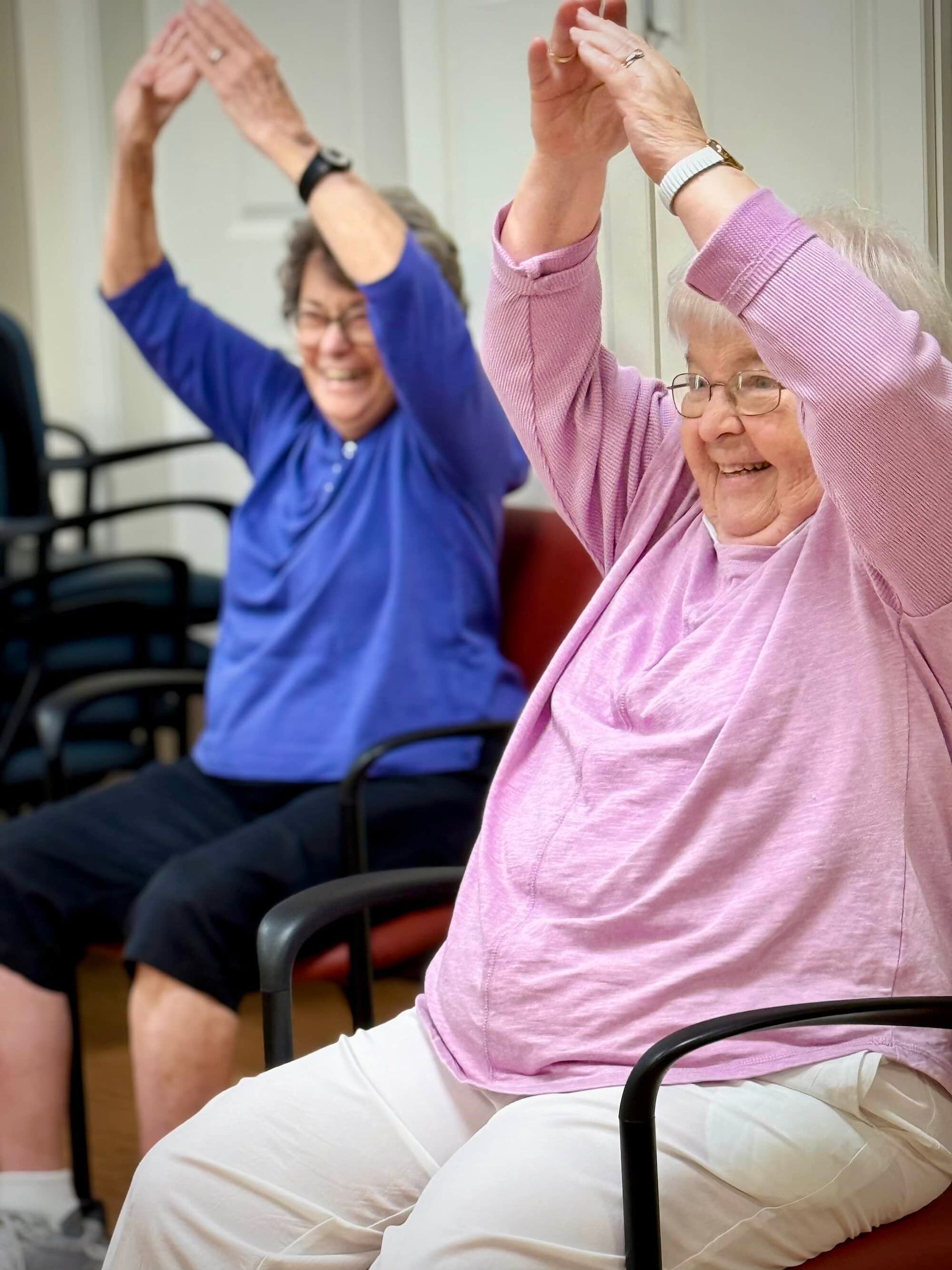 Two elderly women in chairs, smiling and stretching arms overhead. Indoors, wearing casual clothing.
