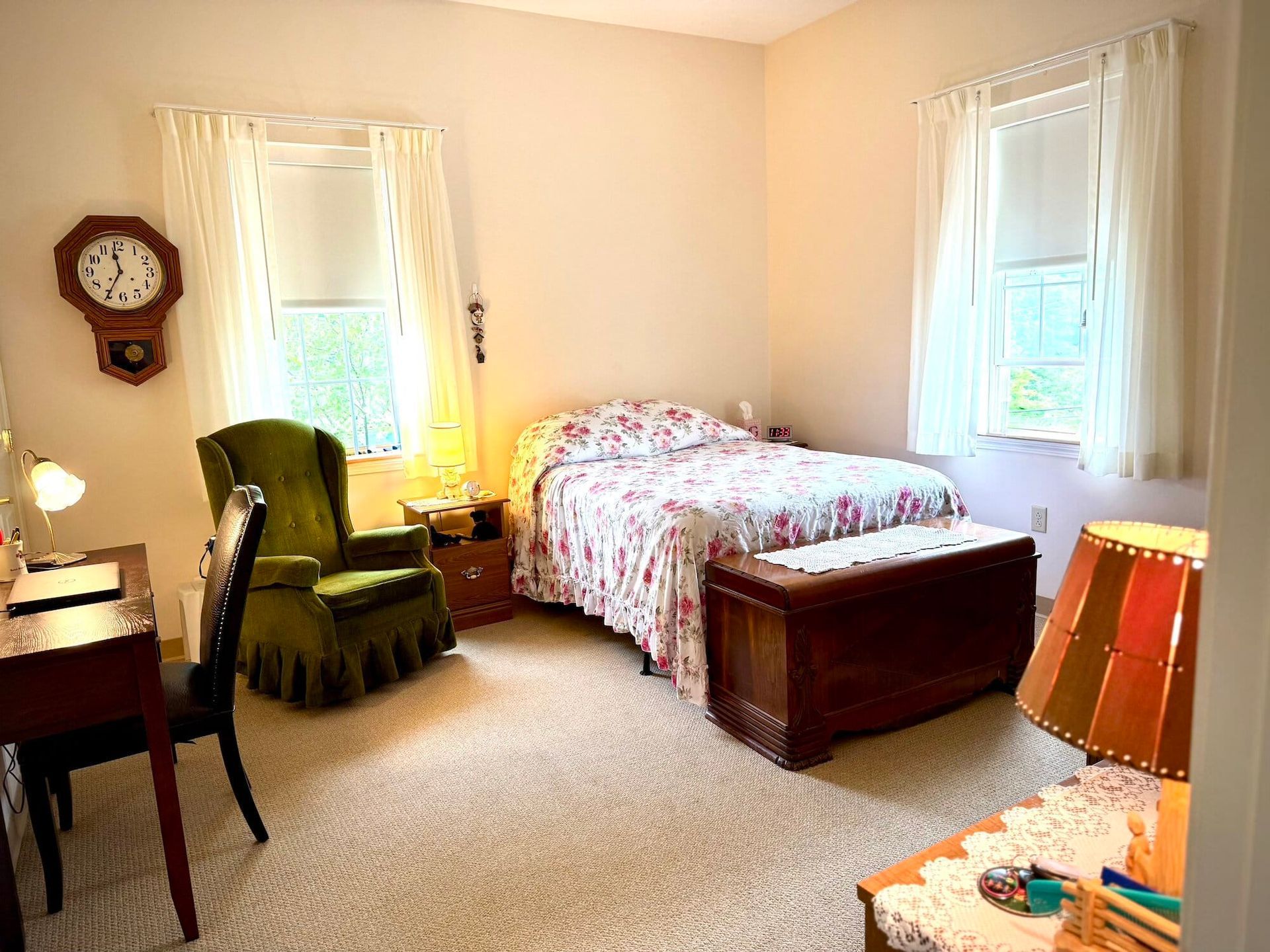 Cozy bedroom with floral bedding, antique clock, green chair, desk, and wooden trunk at the foot of the bed.