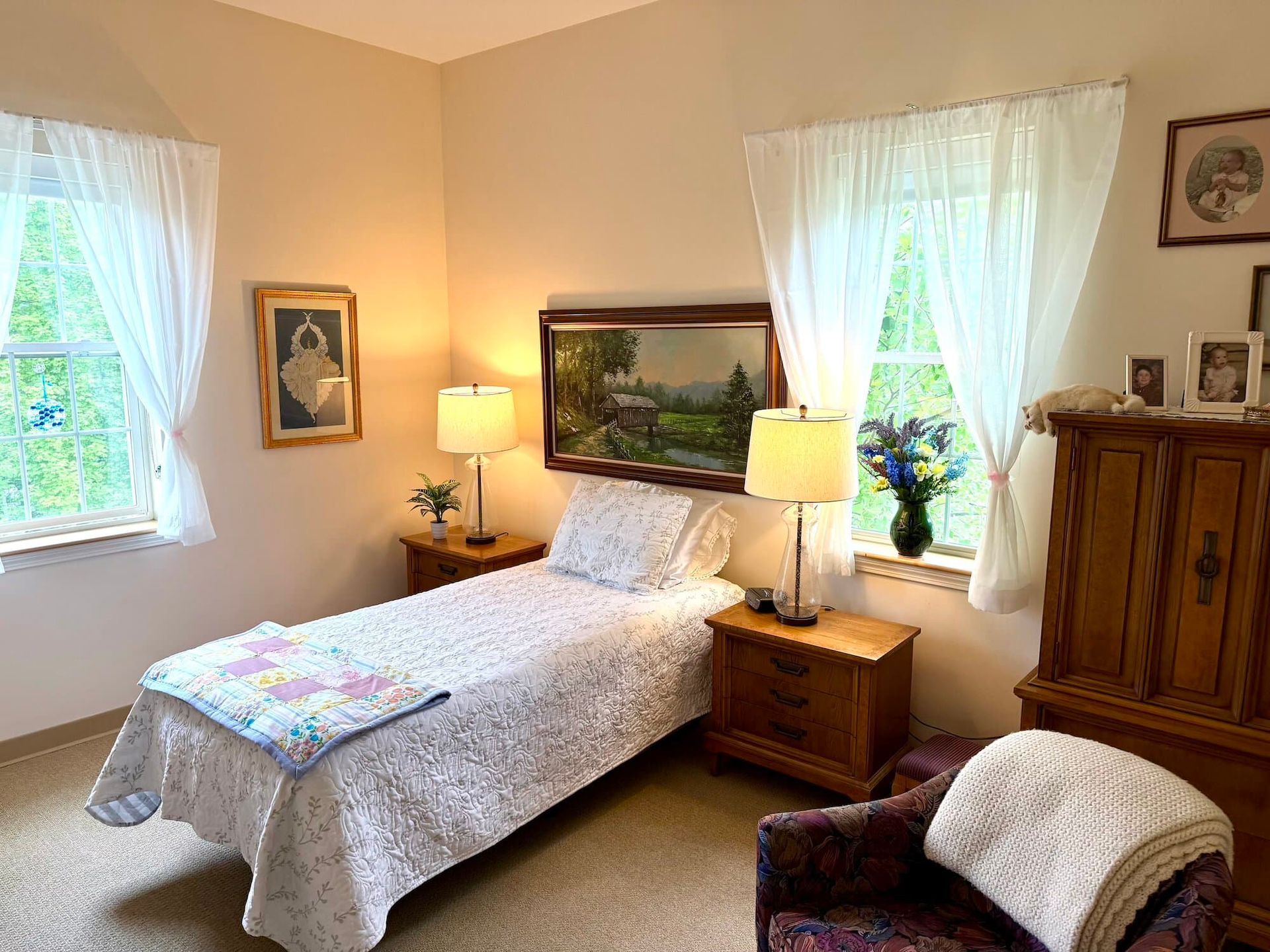 Bedroom with single bed, nightstands, chair, wooden dresser, windows with white curtains, and artwork.