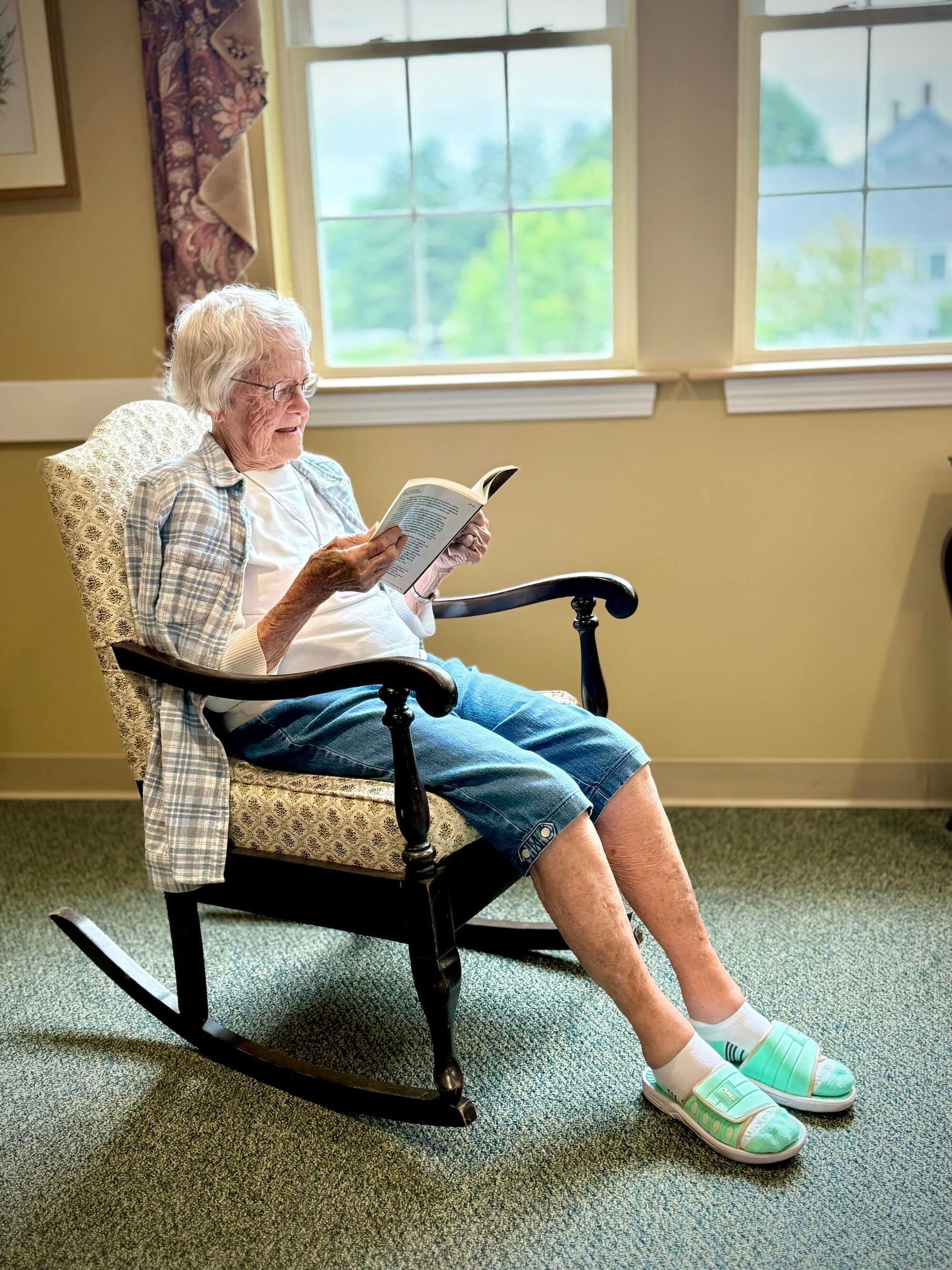 Woman in rocking chair reading book by window.