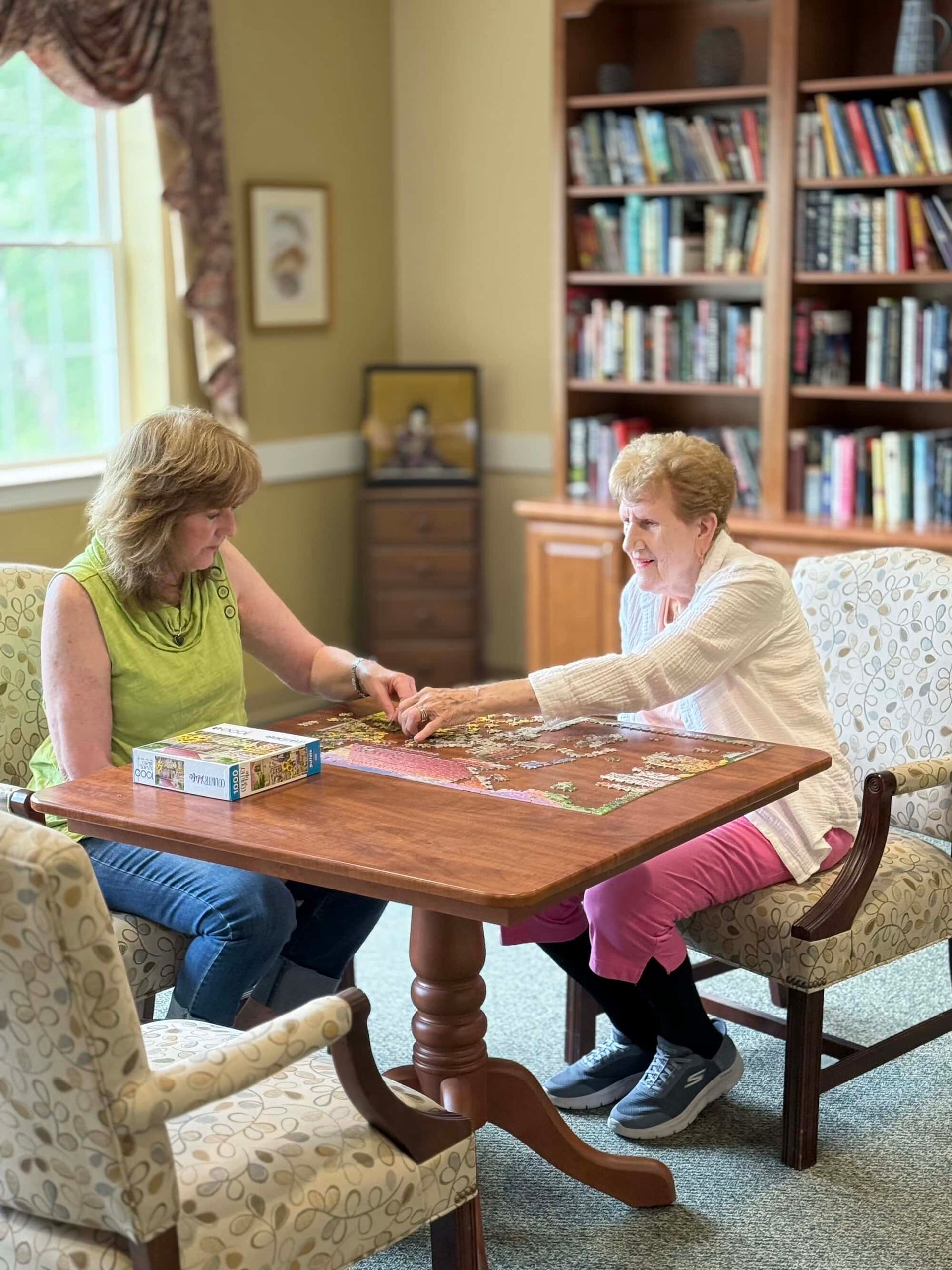 Two women at a table doing a puzzle in a library. One wears pink pants, the other jeans.