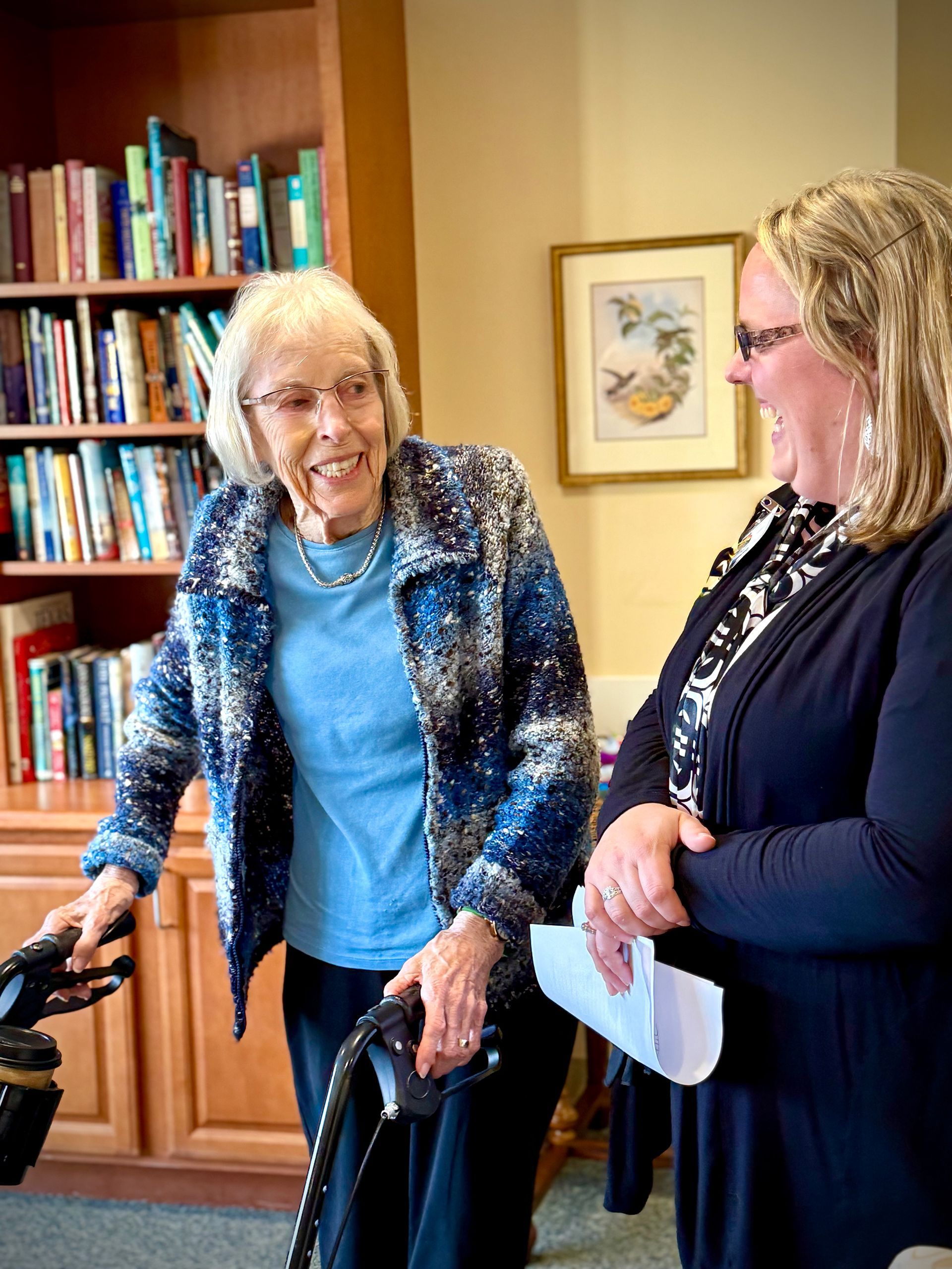 Two women talking indoors near a bookshelf, one using a walker and smiling