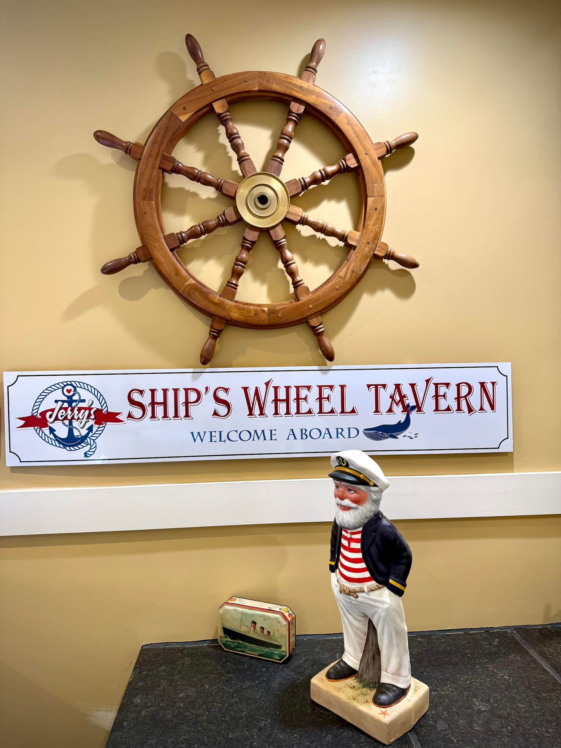 Wooden ship wheel and "Ship's Wheel Tavern" sign above a captain figurine on a counter.