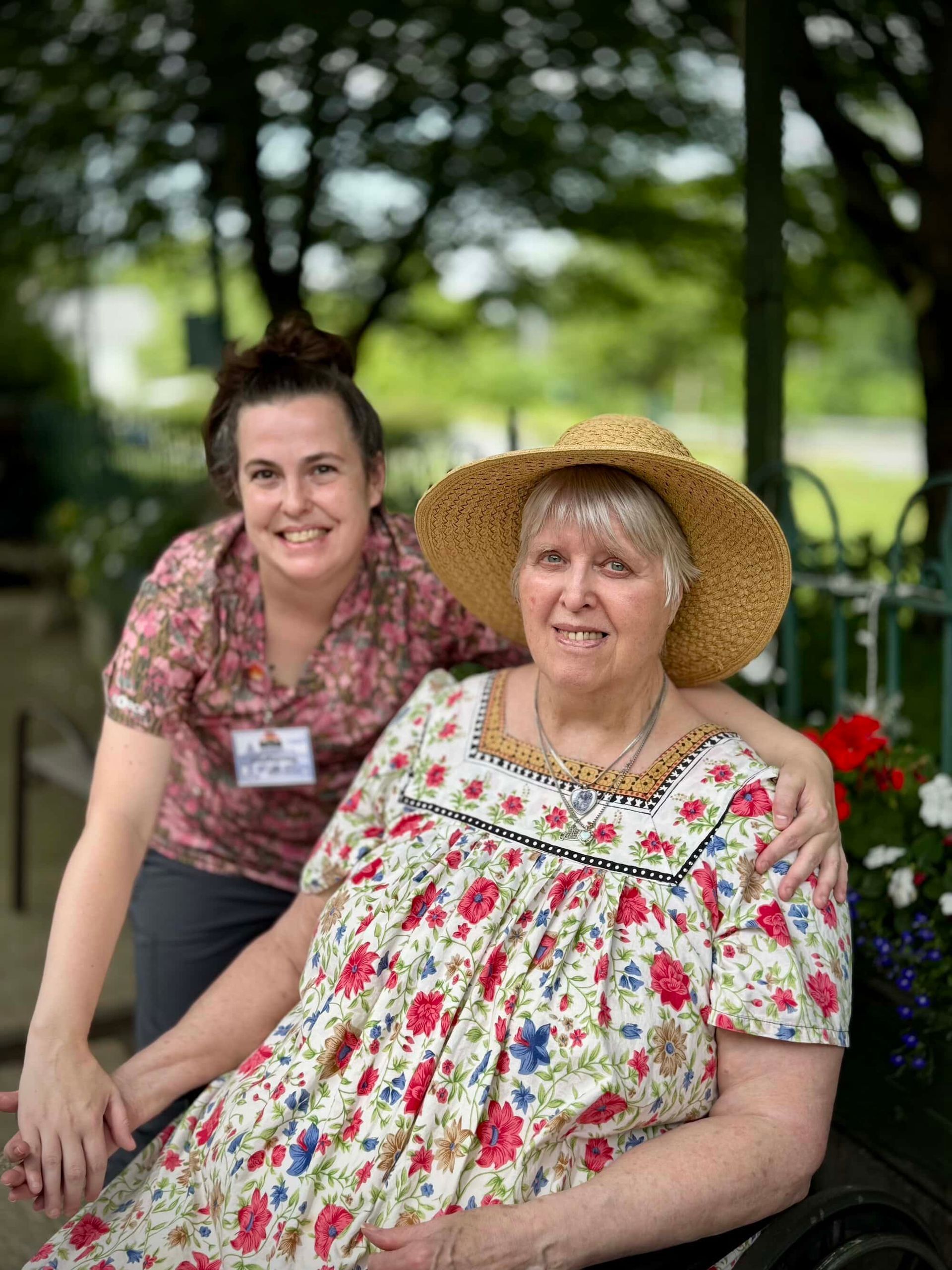 Woman in straw hat smiles with arm around another woman, both outdoors near flowers.