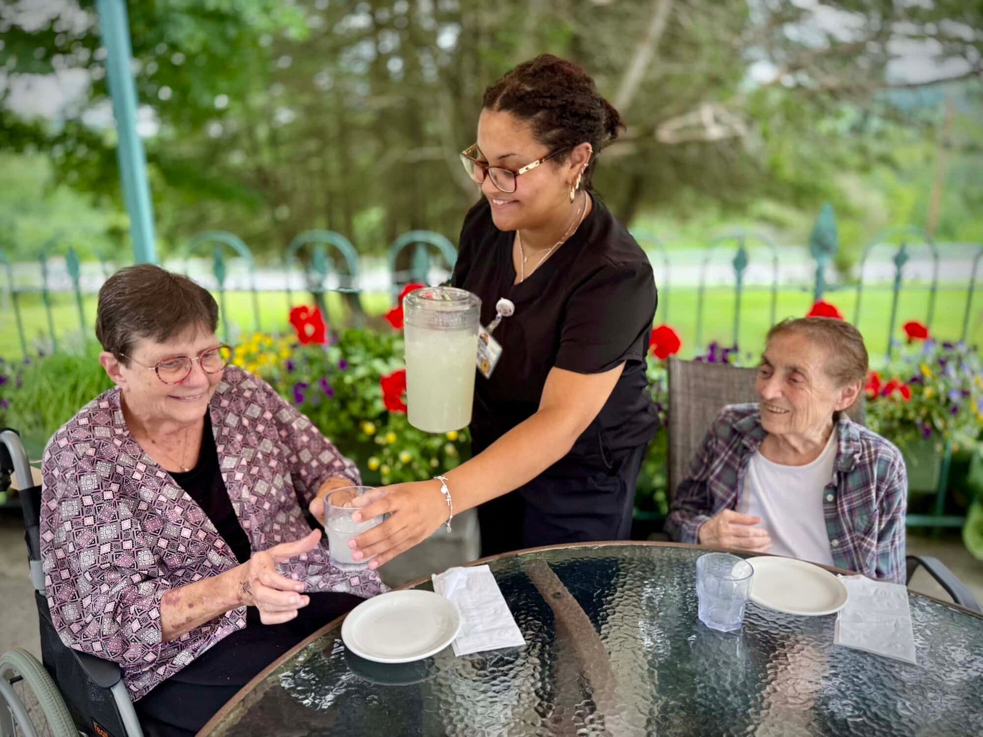 Caregiver pours drinks for two people seated at an outdoor table.