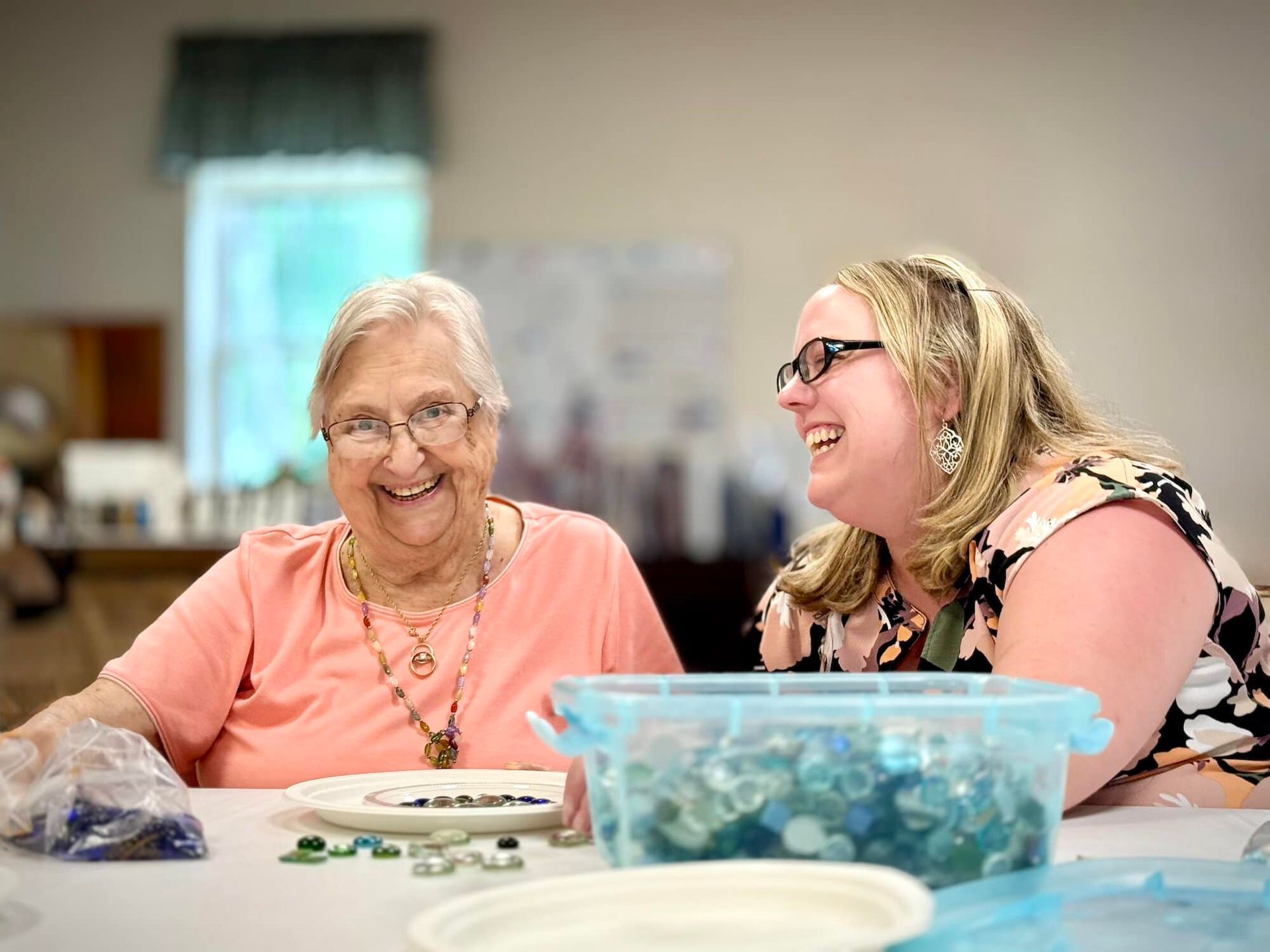 Two women smiling at a table, craft supplies. Older woman in peach, younger woman in glasses laughing.