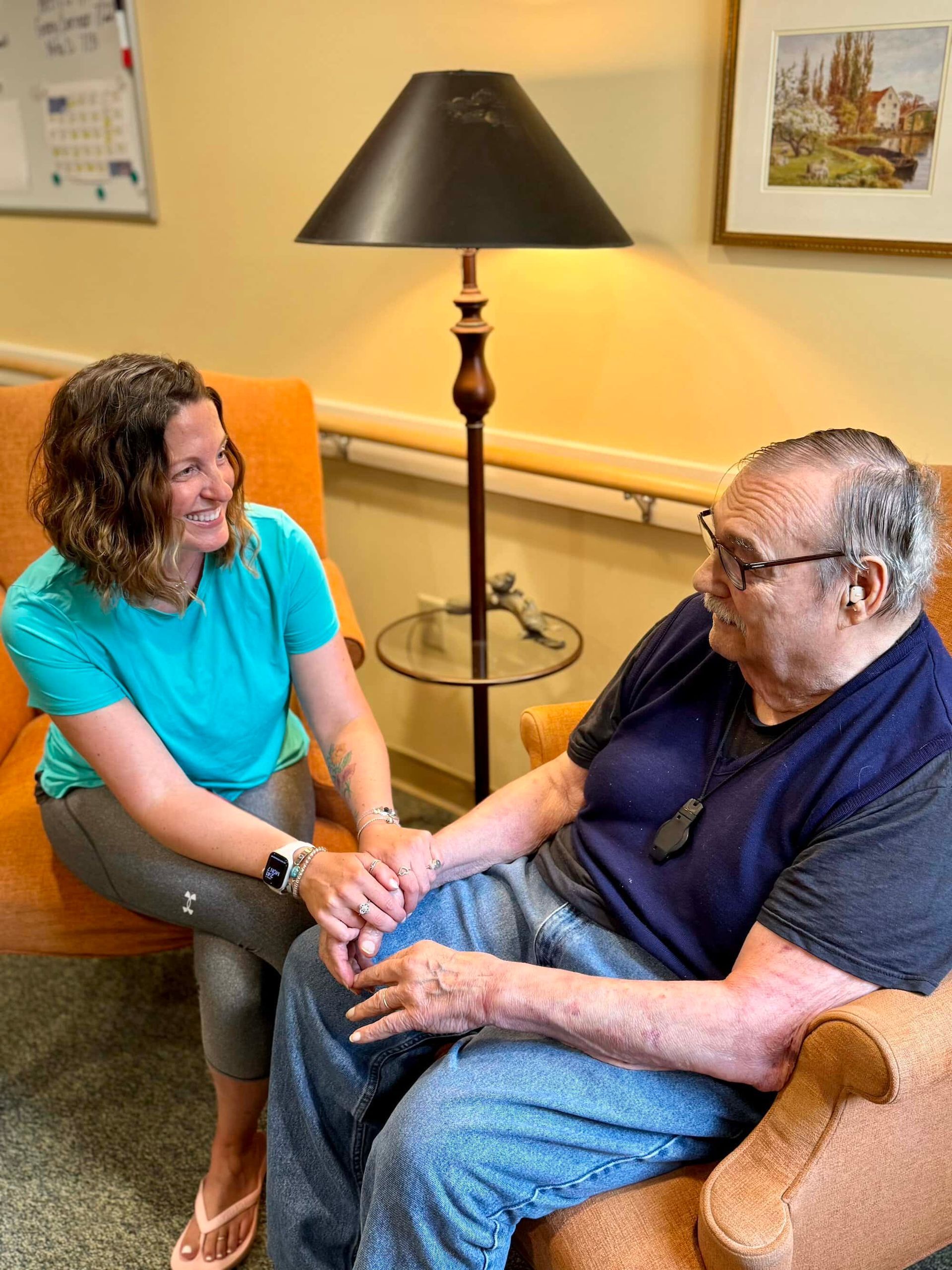 Woman holding hands with older person, smiling, seated in chairs, lamp in the background.