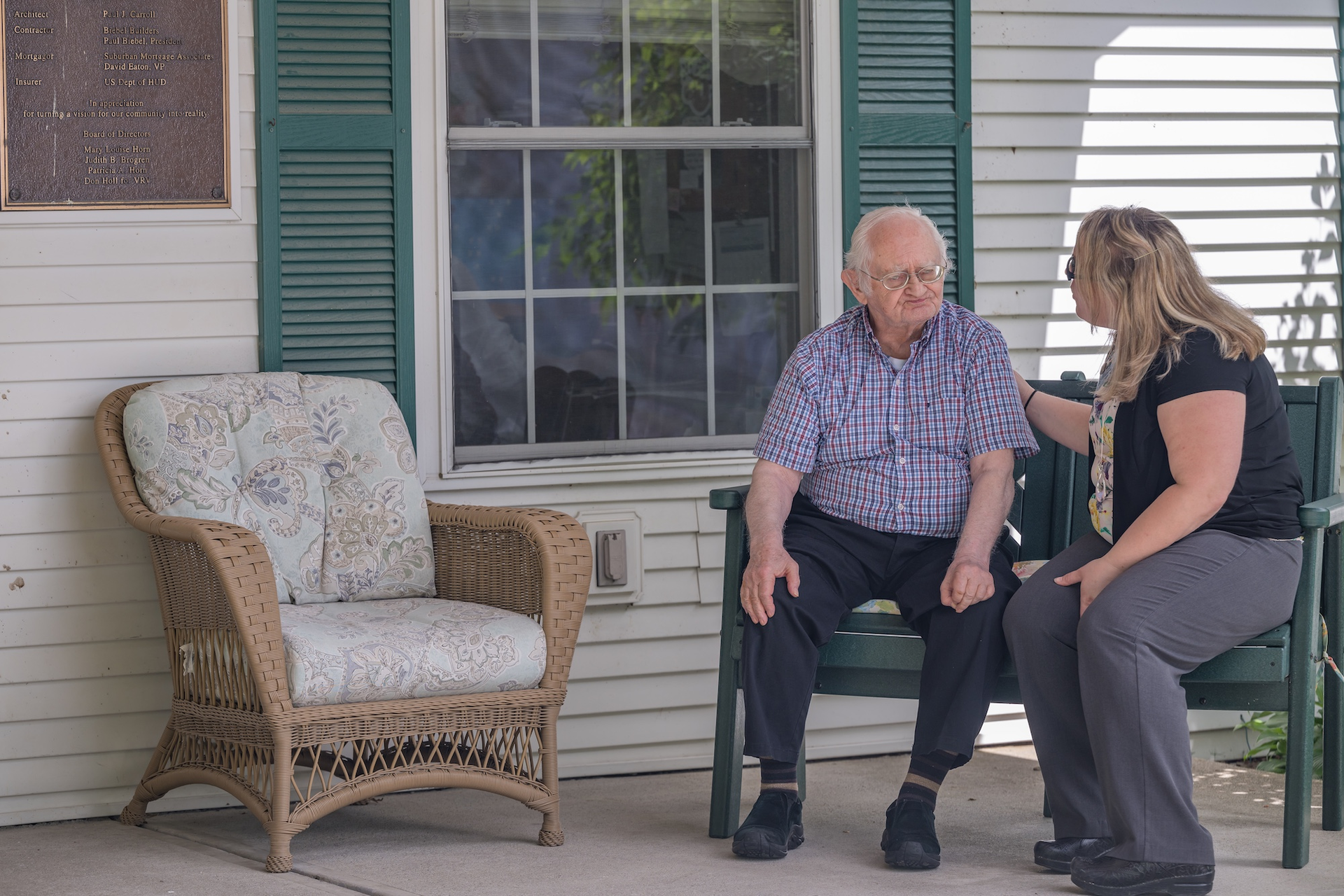 Elderly man sits on a bench, talking to a person. They are on a porch.