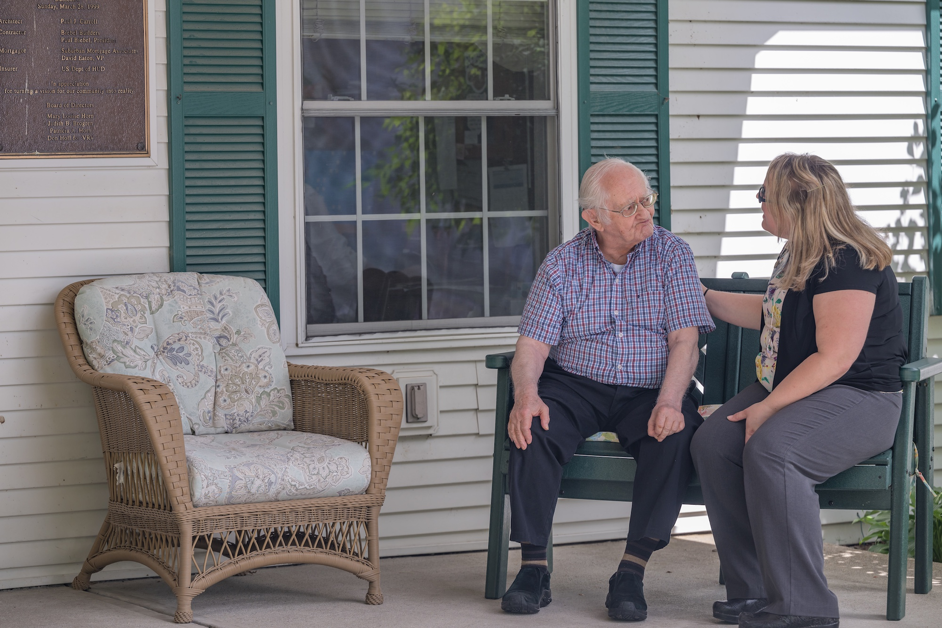 Man and woman seated on porch bench, woman has arm on man's shoulder. Natural light.