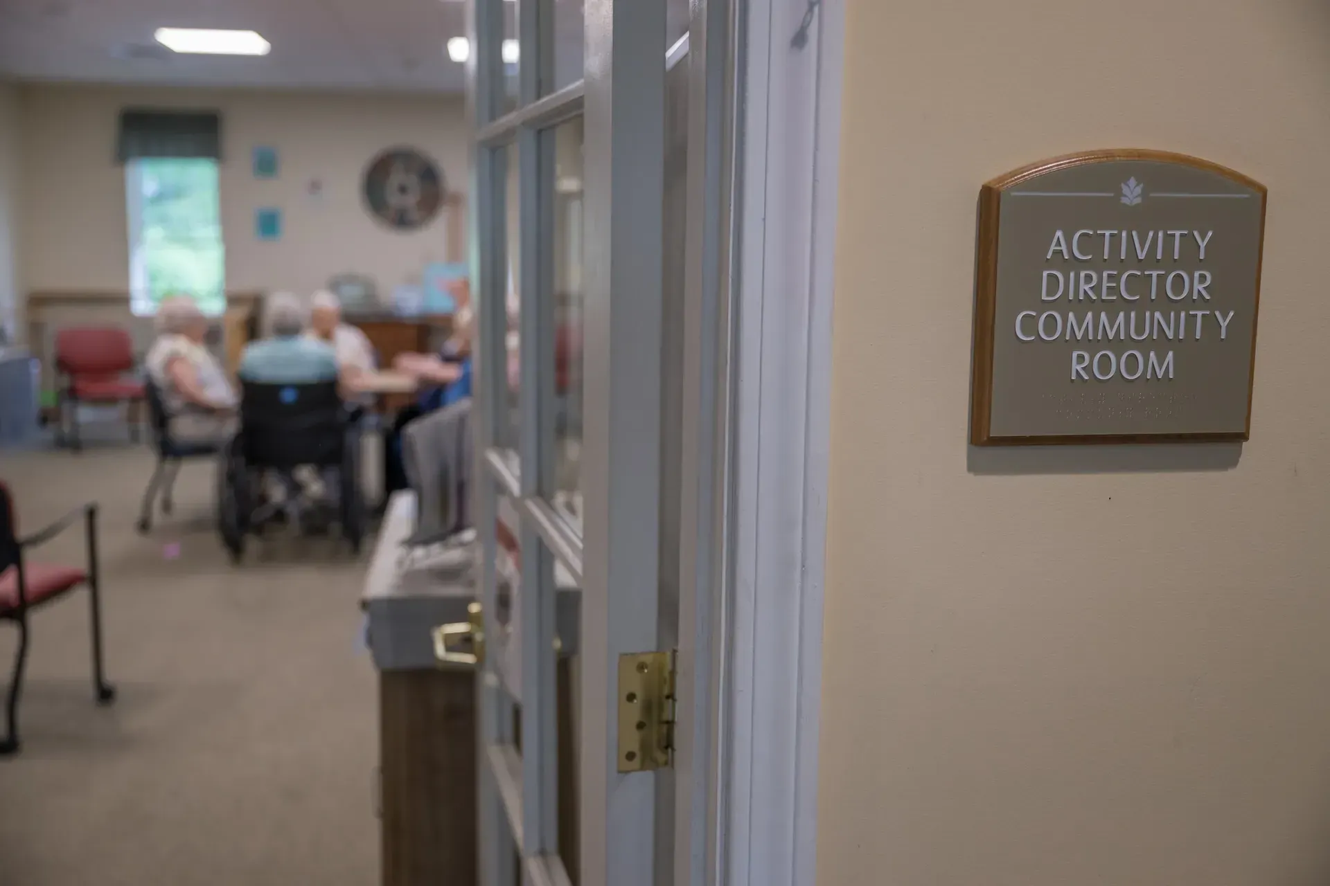 Doorway into Activity Director/Community Room, people seated at table in the background. Sign is on the wall.