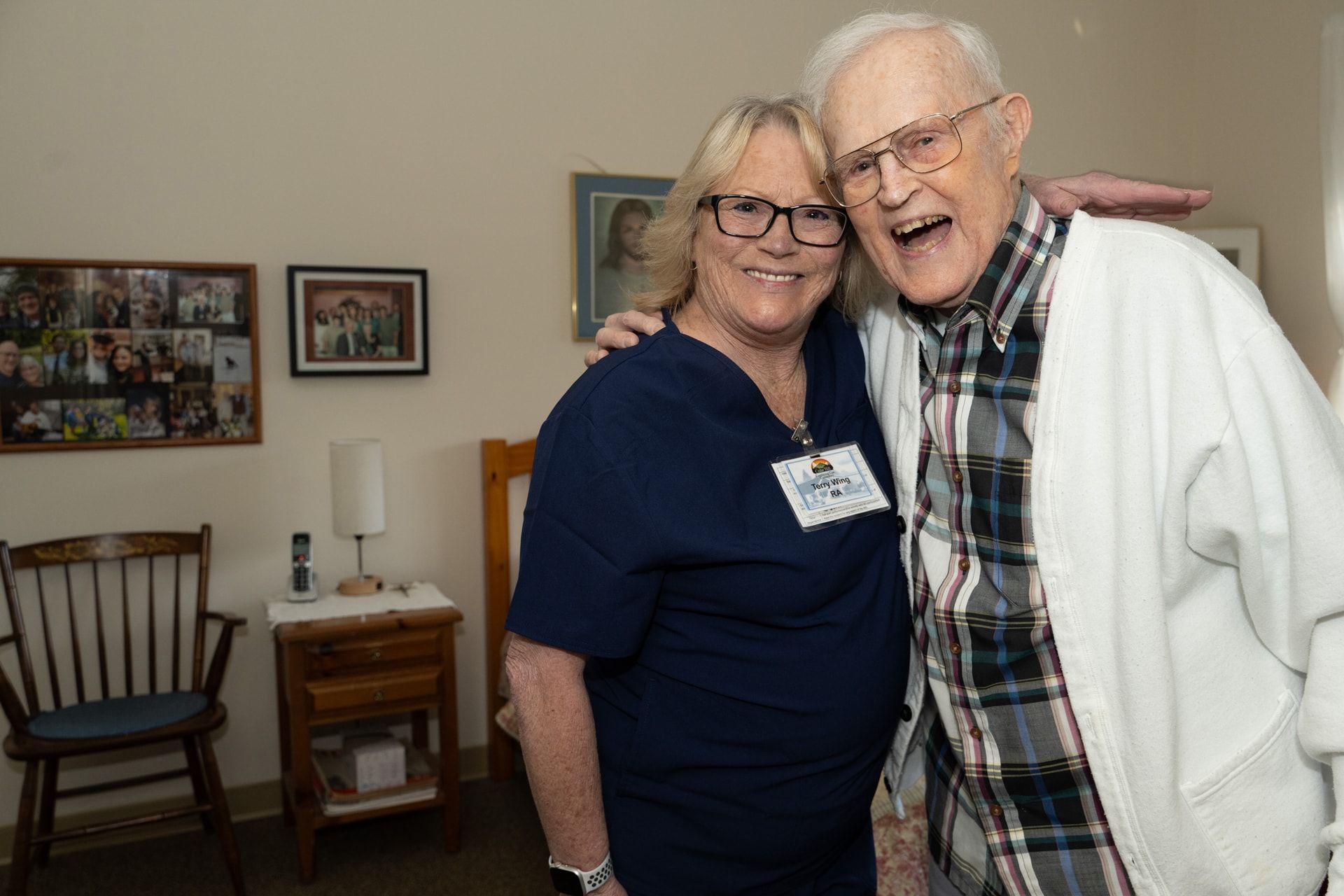 Woman in blue scrubs embraces an elderly man. They smile in a room with furniture and framed photos.