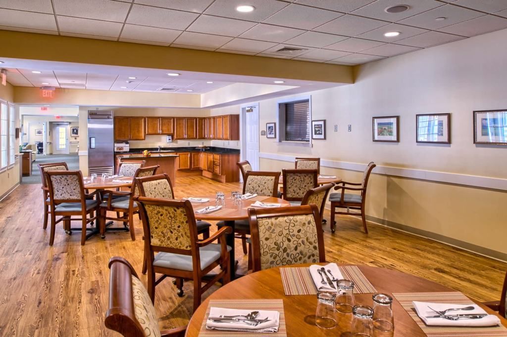 Dining area with round tables, chairs, and a kitchen in the background. Light wood floors and neutral walls.