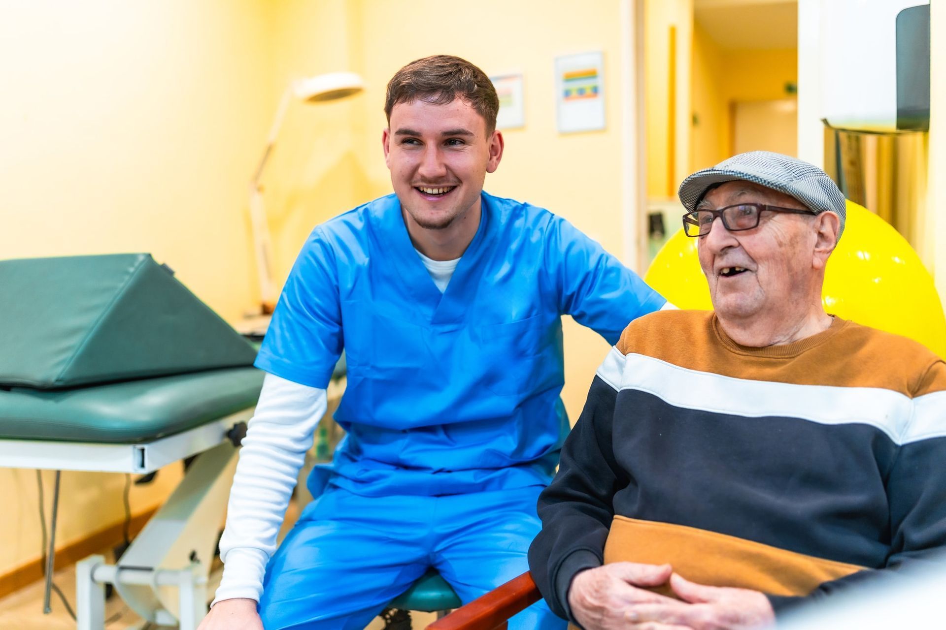 Smiling healthcare worker in blue scrubs with an older person in a room, the patient sitting and wearing a cap.