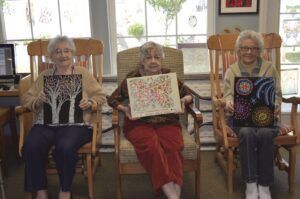 Three elderly women in rocking chairs holding their artwork; indoor setting with windows.