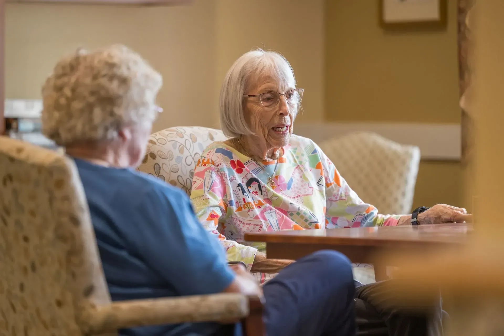 Two older women seated at a table, conversing indoors. One wears glasses and a patterned shirt.