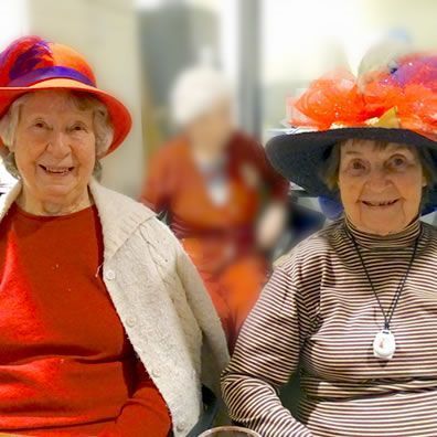 Two older women smiling, wearing decorative hats. One in red, one in stripes, with a blurred woman behind them.