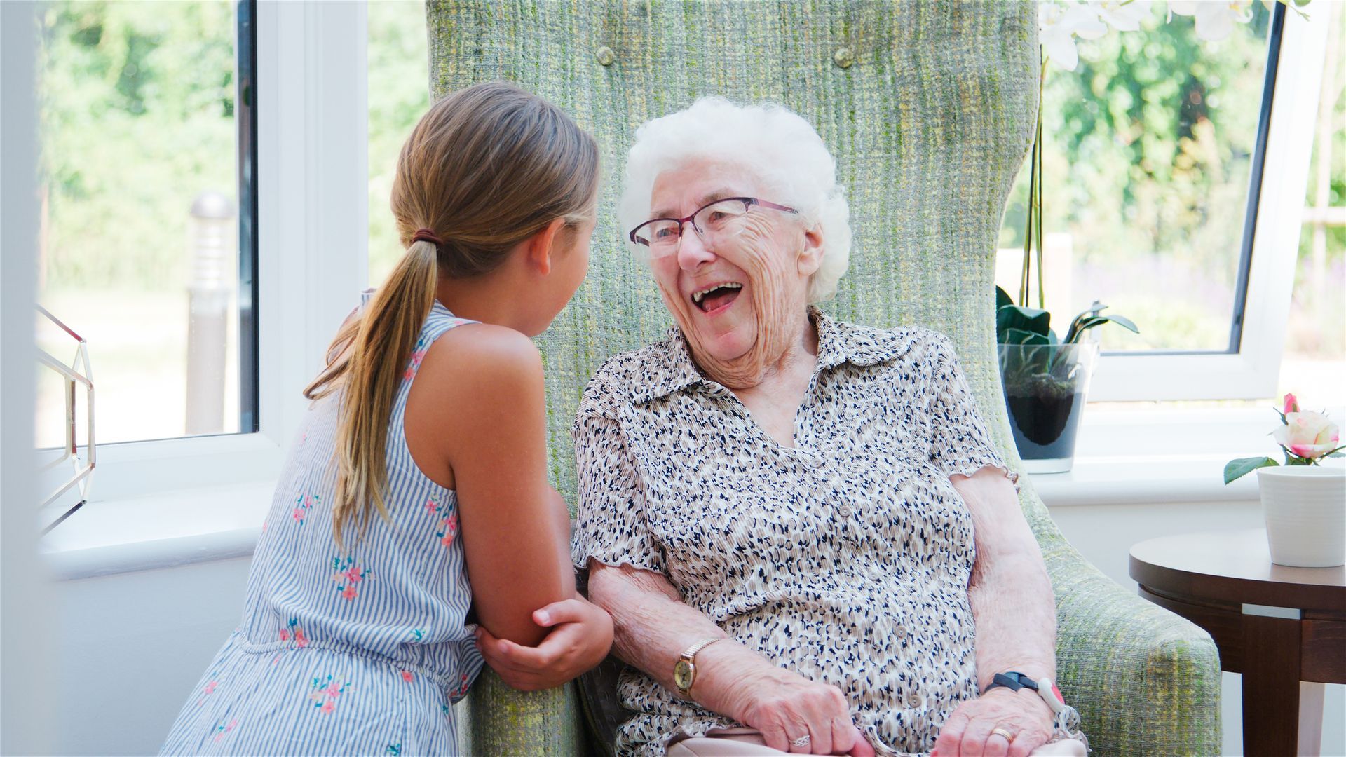 A child and an older adult laugh while facing each other, seated together in a sunlit room.