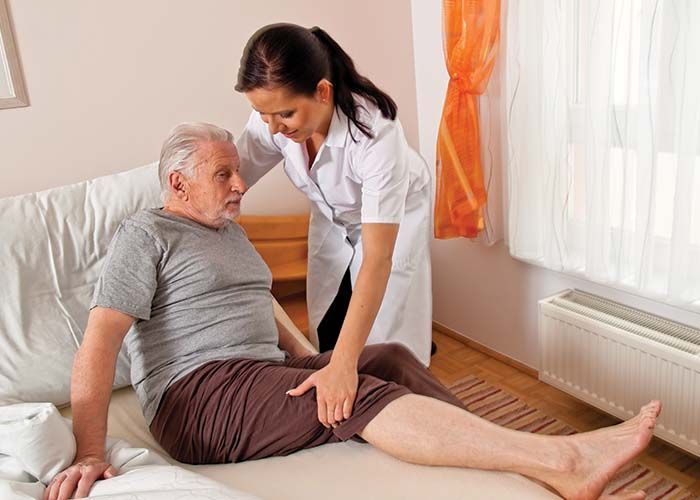 Caregiver assisting a person sitting on a bed; indoor setting; touching leg.