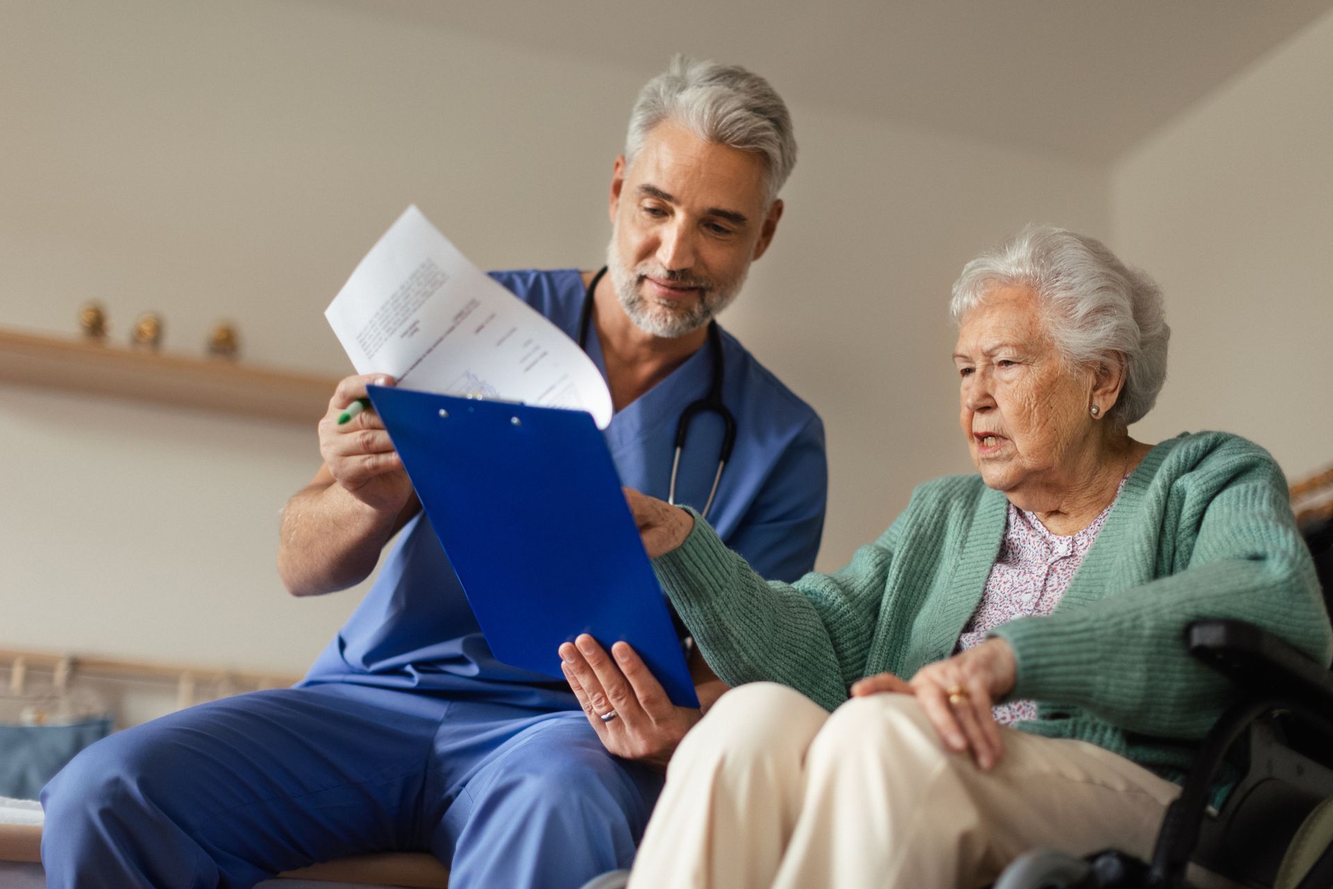 Man in blue scrubs shows document on clipboard to older person in a wheelchair.