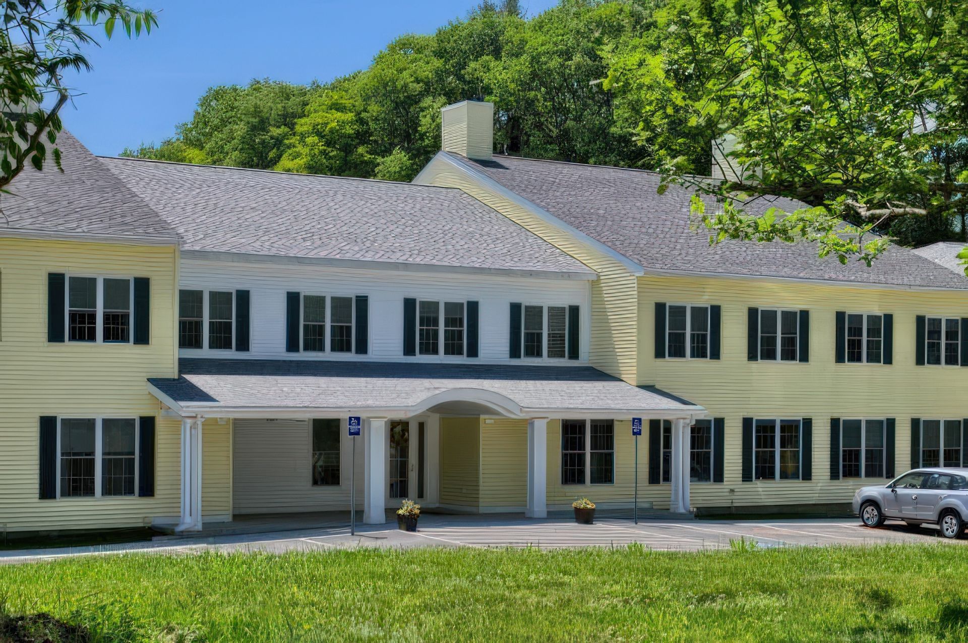 Yellow building with gray roof and green shutters, in a grassy area, with a parked car.