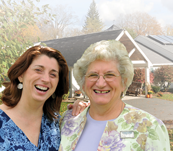 Two women smiling in front of a building with trees in the background.