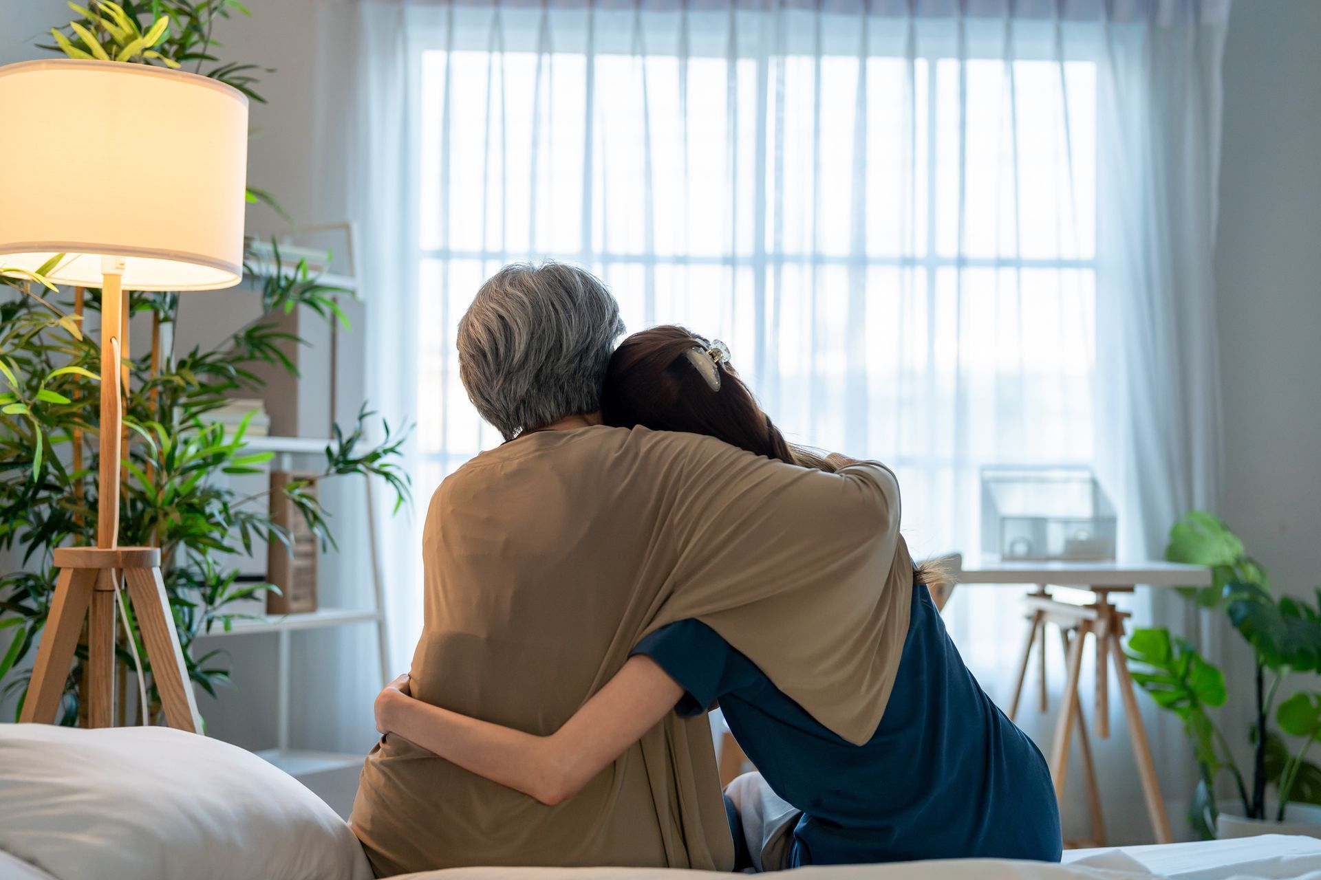 Woman hugging another woman, both looking out window. Room with lamp, plants, and a bed.