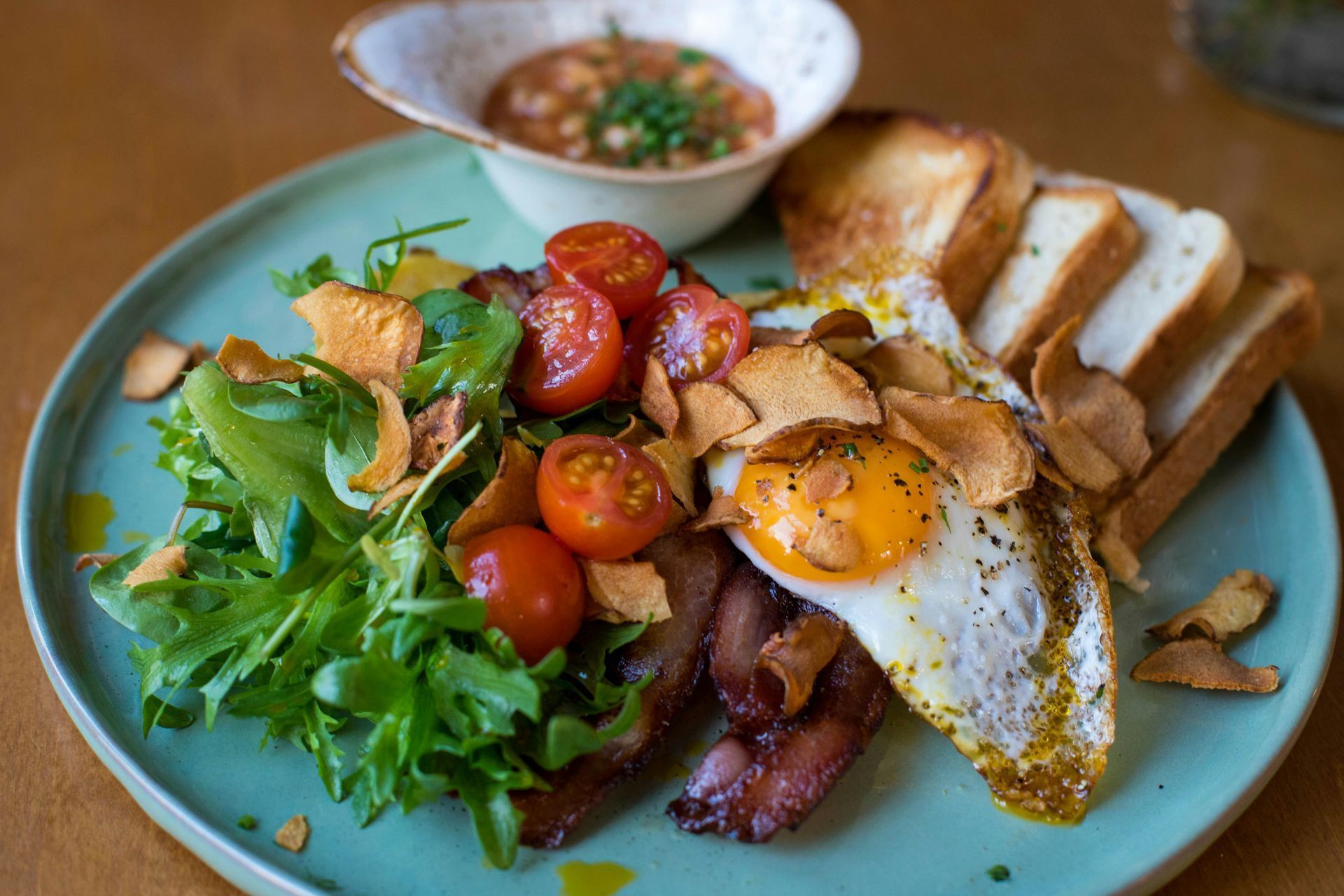 Breakfast plate with fried egg, bacon, tomatoes, salad, toast, and bean dip.