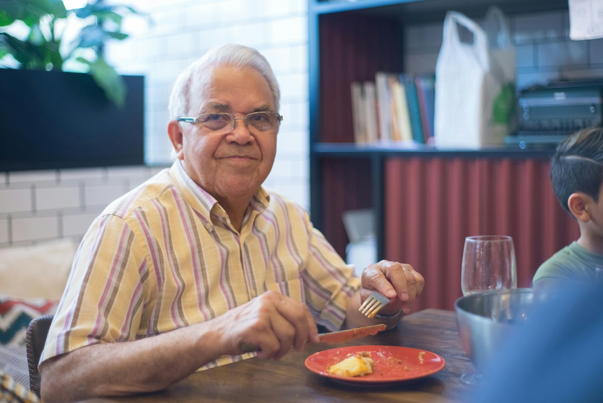 Man with glasses seated at a table, holding fork and knife, red plate with food.