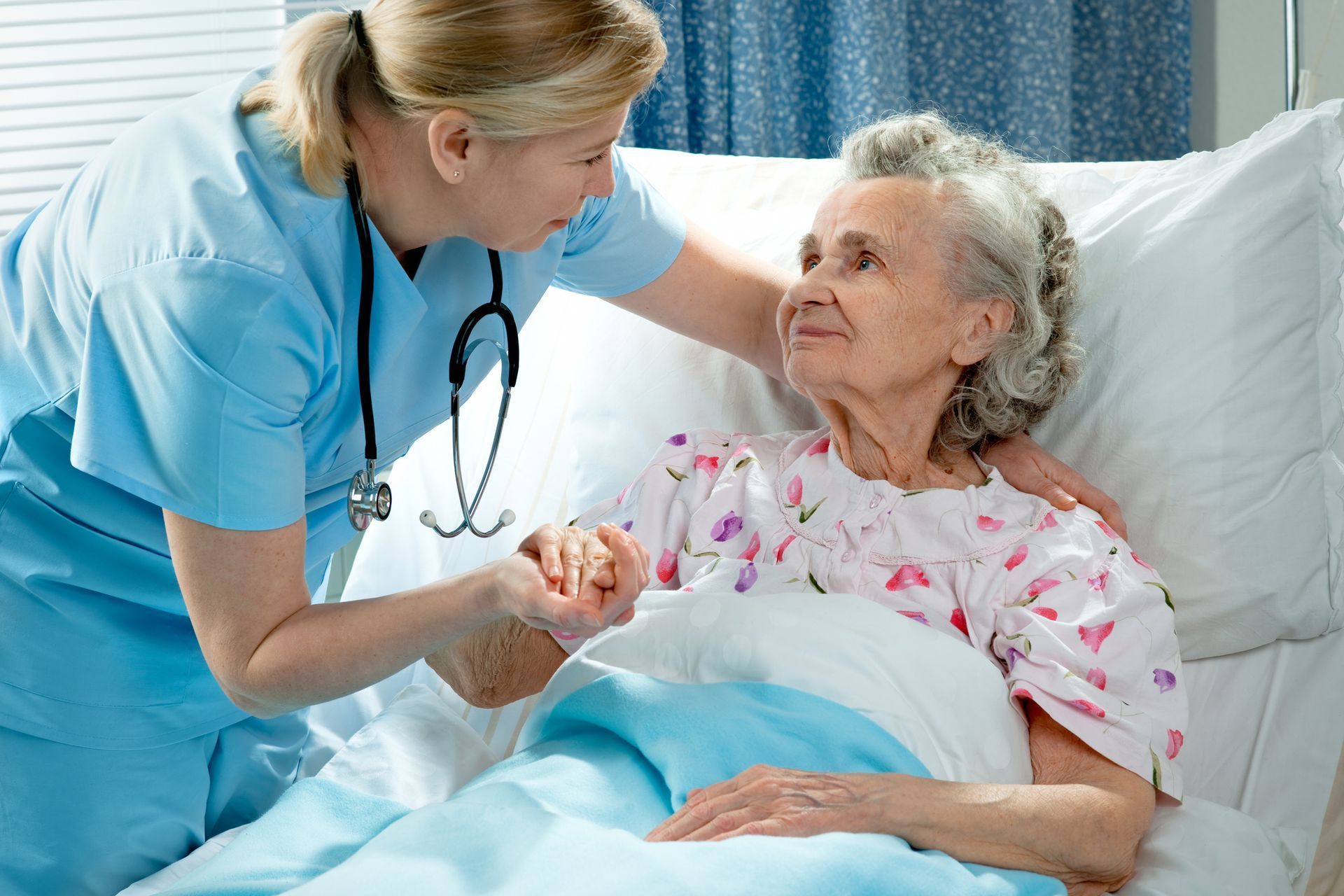 Nurse in scrubs comforting an elderly person in bed, holding hands. Hospital setting, gentle expression.