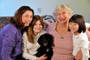Four smiling women and a black dog pose indoors.