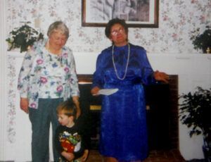 Two women and a child pose near a fireplace; one woman in blue holds paper, other in floral top.
