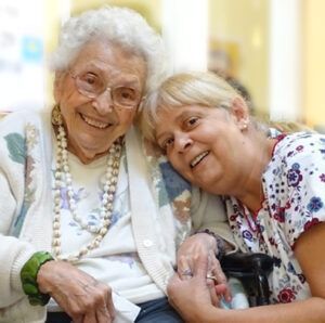 An elderly woman smiles with a younger woman, who rests her head on the older woman's shoulder. Both are smiling.