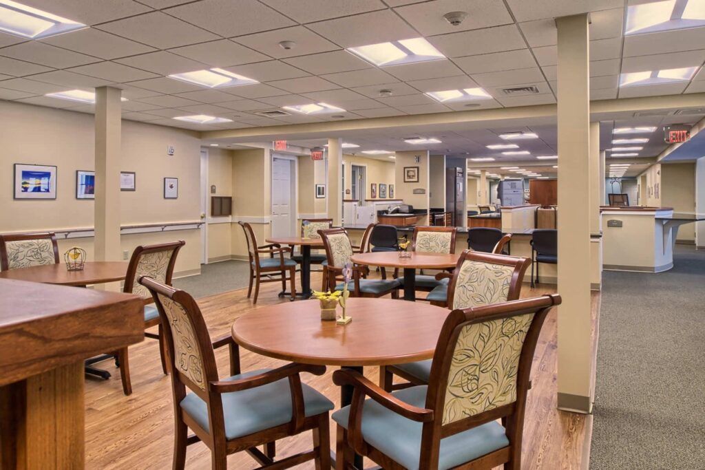 Empty dining area in a senior living facility with round tables, patterned chairs, and neutral tones.