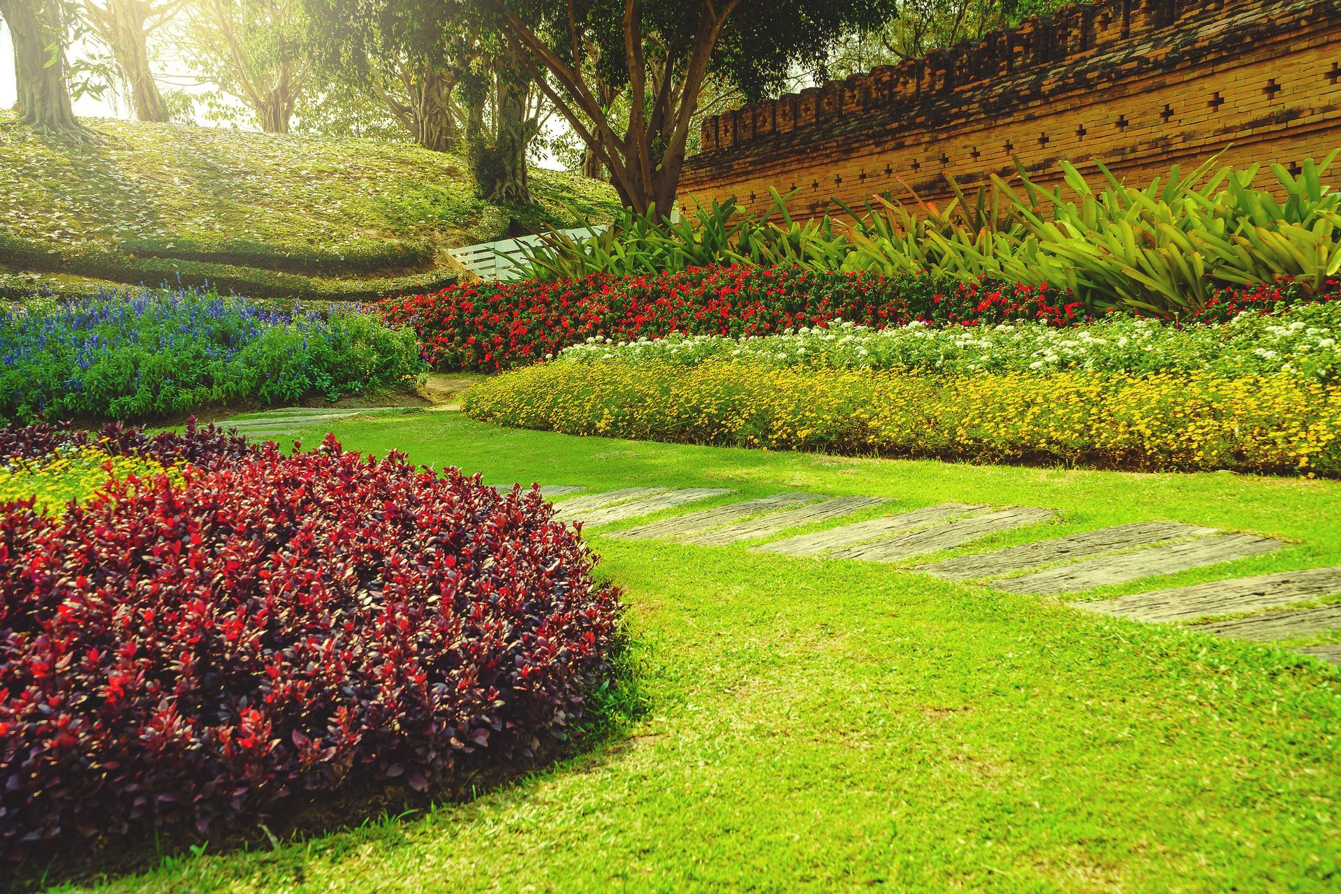 Colorful tiered garden with stone path and red, yellow, and green flowers, brick wall, and green lawn.