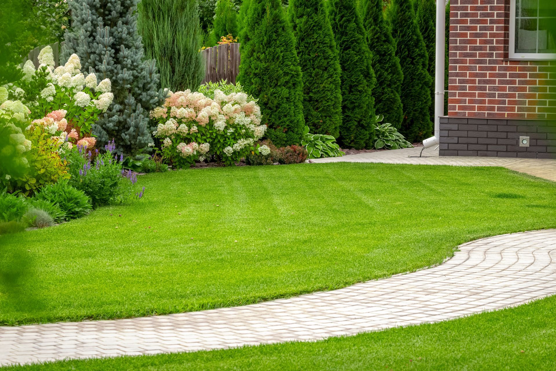 Lush green lawn with a curved stone pathway, bordered by colorful flowering shrubs and evergreen trees near a brick house.