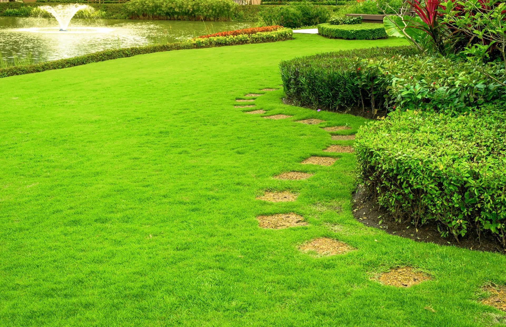 Lush green lawn with a stone path winding past manicured bushes towards a pond with a fountain.