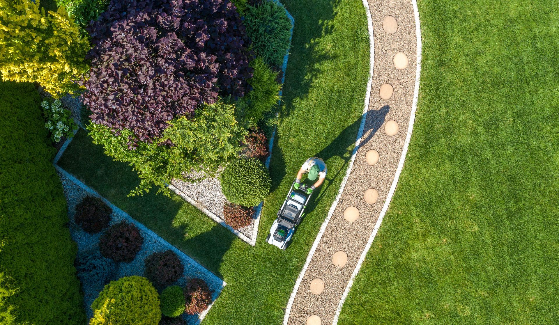 Overhead view of a garden with a winding stone path, green lawn, flower beds, and a lawnmower.