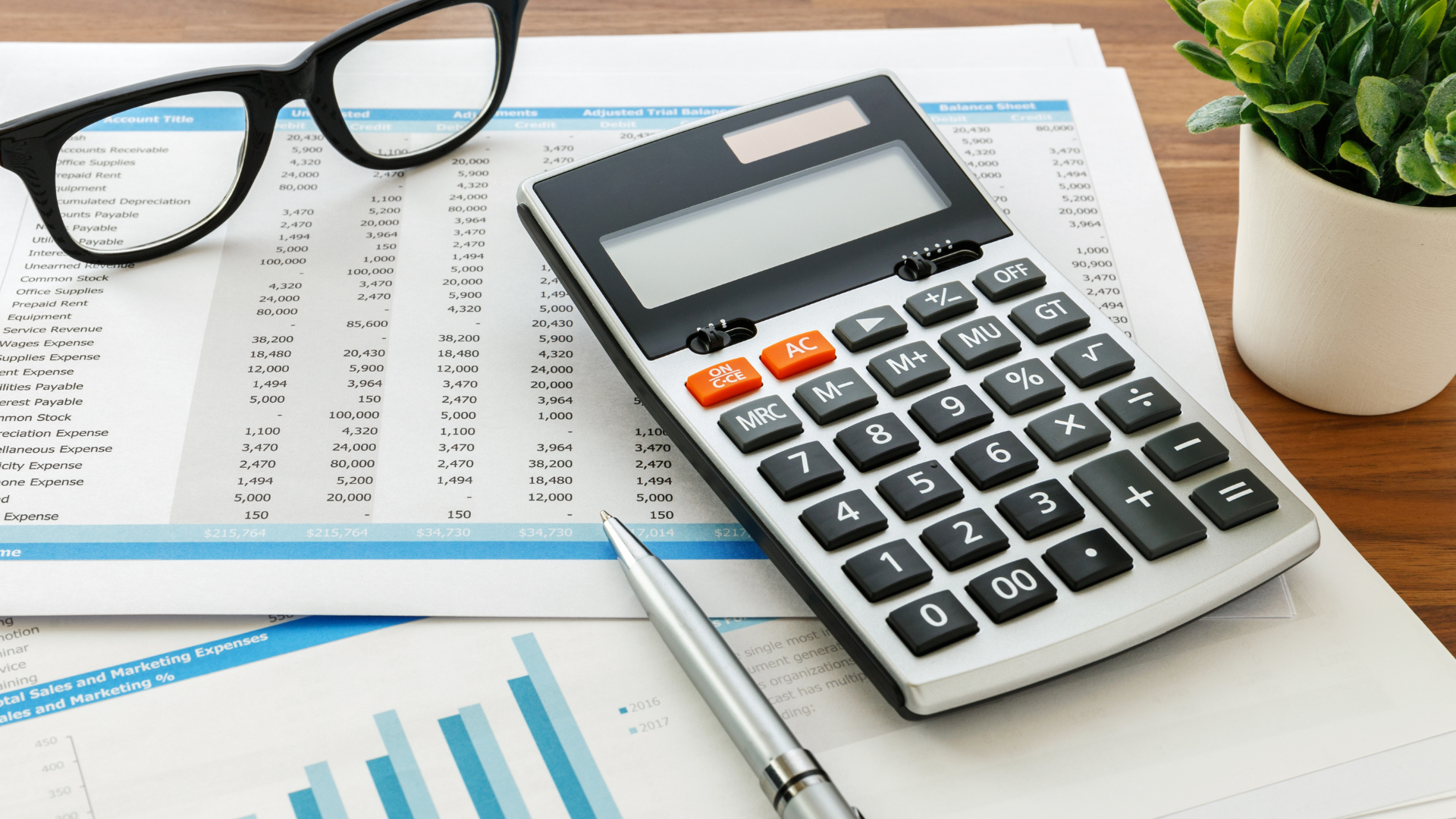 A calculator , pen , glasses and a potted plant are on a table.