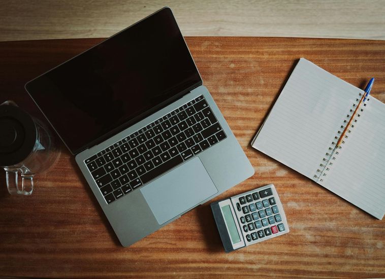 A laptop computer is sitting on a wooden desk next to a notebook and a calculator.