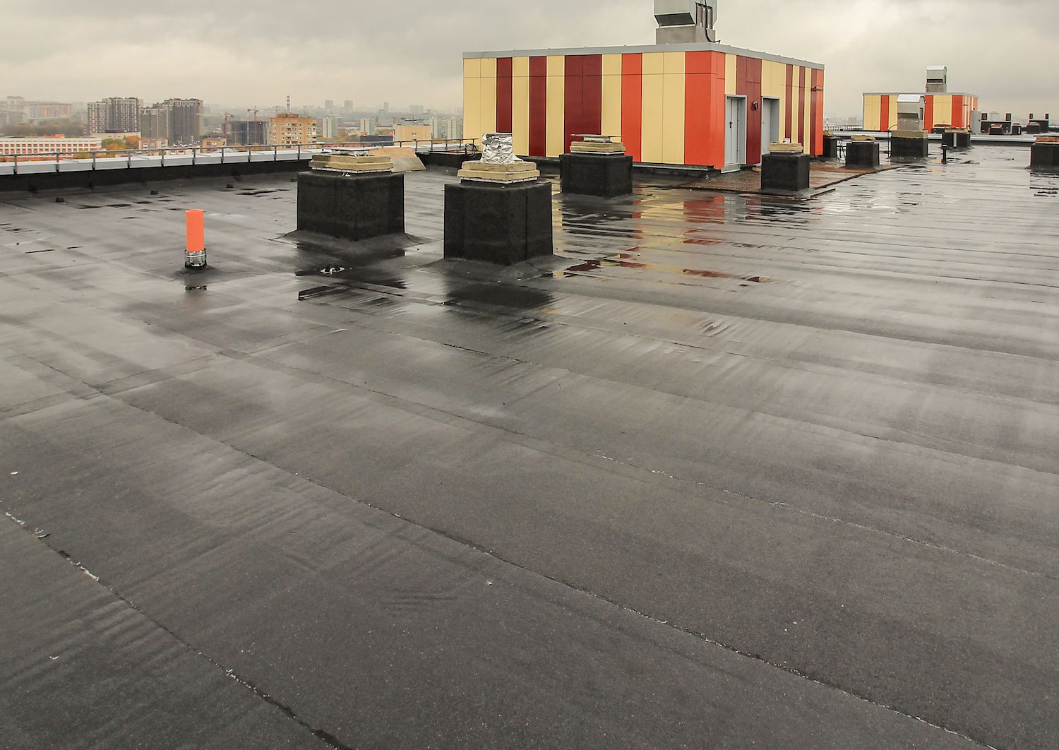A wet, flat, black tar roof with several concrete blocks, against a cityscape and a utility structure.