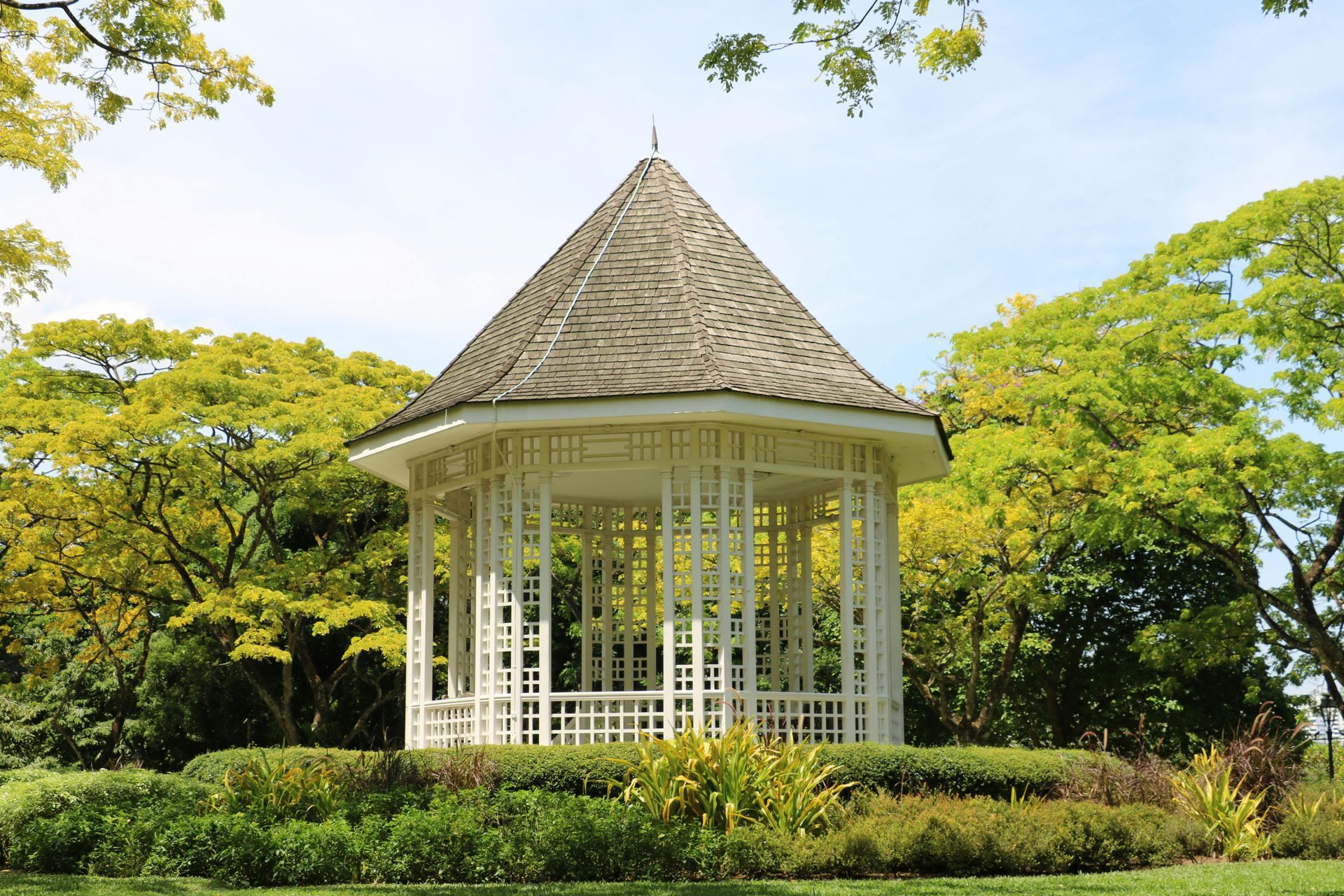 White gazebo with a gray roof in a lush green garden under a sunny sky.