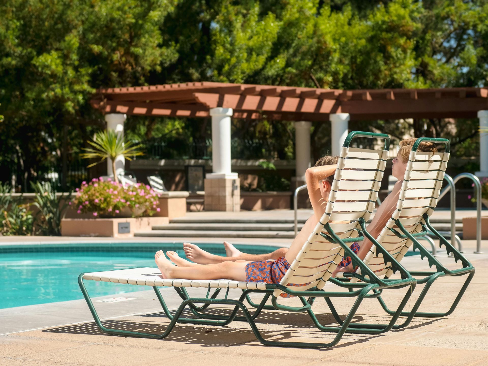Two people sunbathing on lounge chairs by a pool, under a pergola.